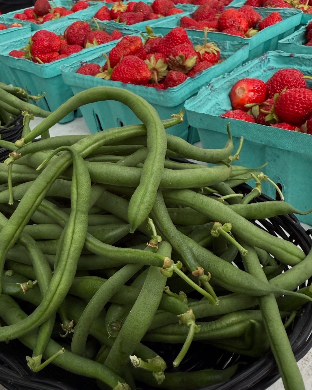 Fresh green beans in a basket with boxes of strawberries in the background at a market.