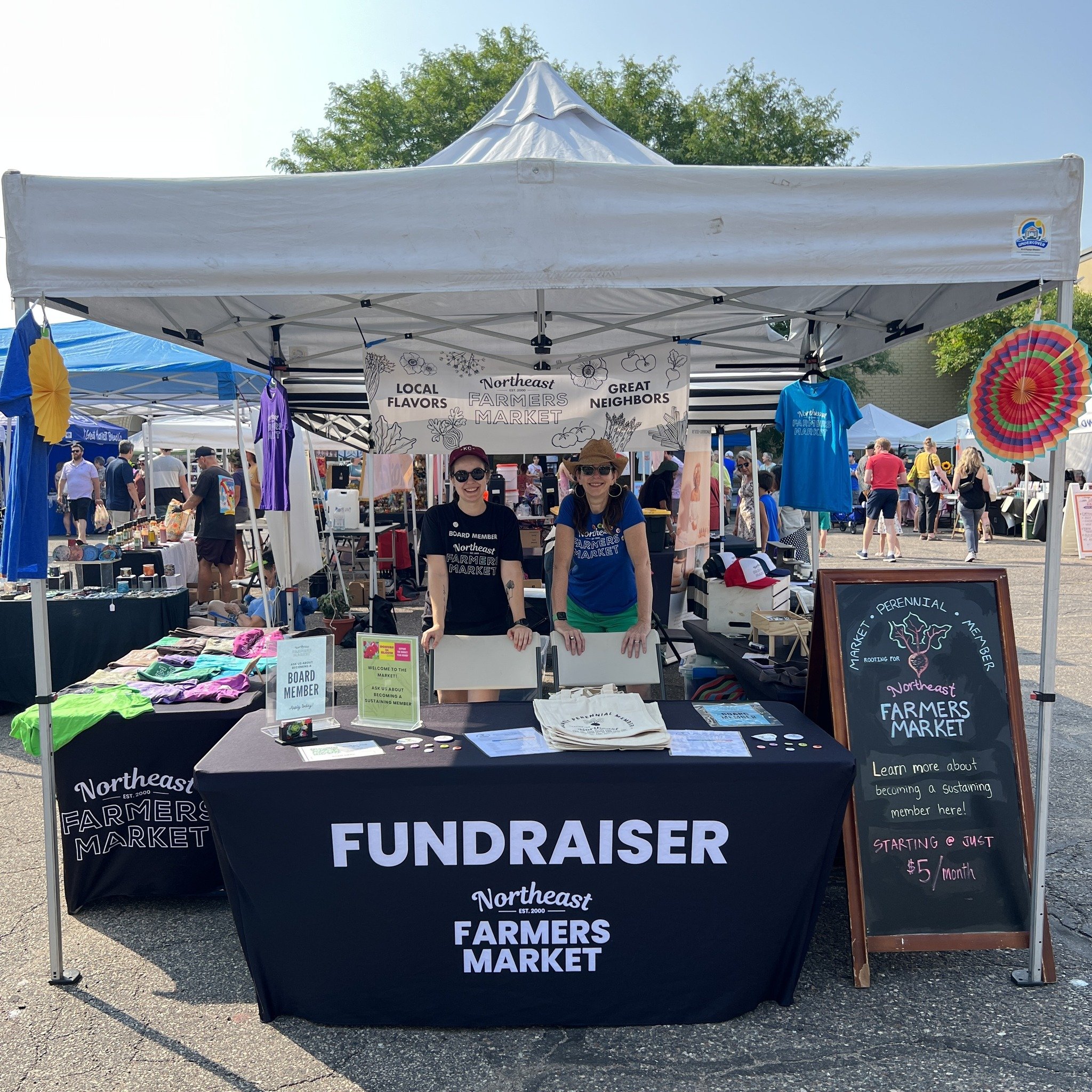 A white canopy tent at a farmers market with a sign that says 'Northeast Farmers Market'. There are two women standing behind a table with various items, and people walking in the background. The sky is clear and blue, and there is a blackboard sign on the right advertising a fundraiser.