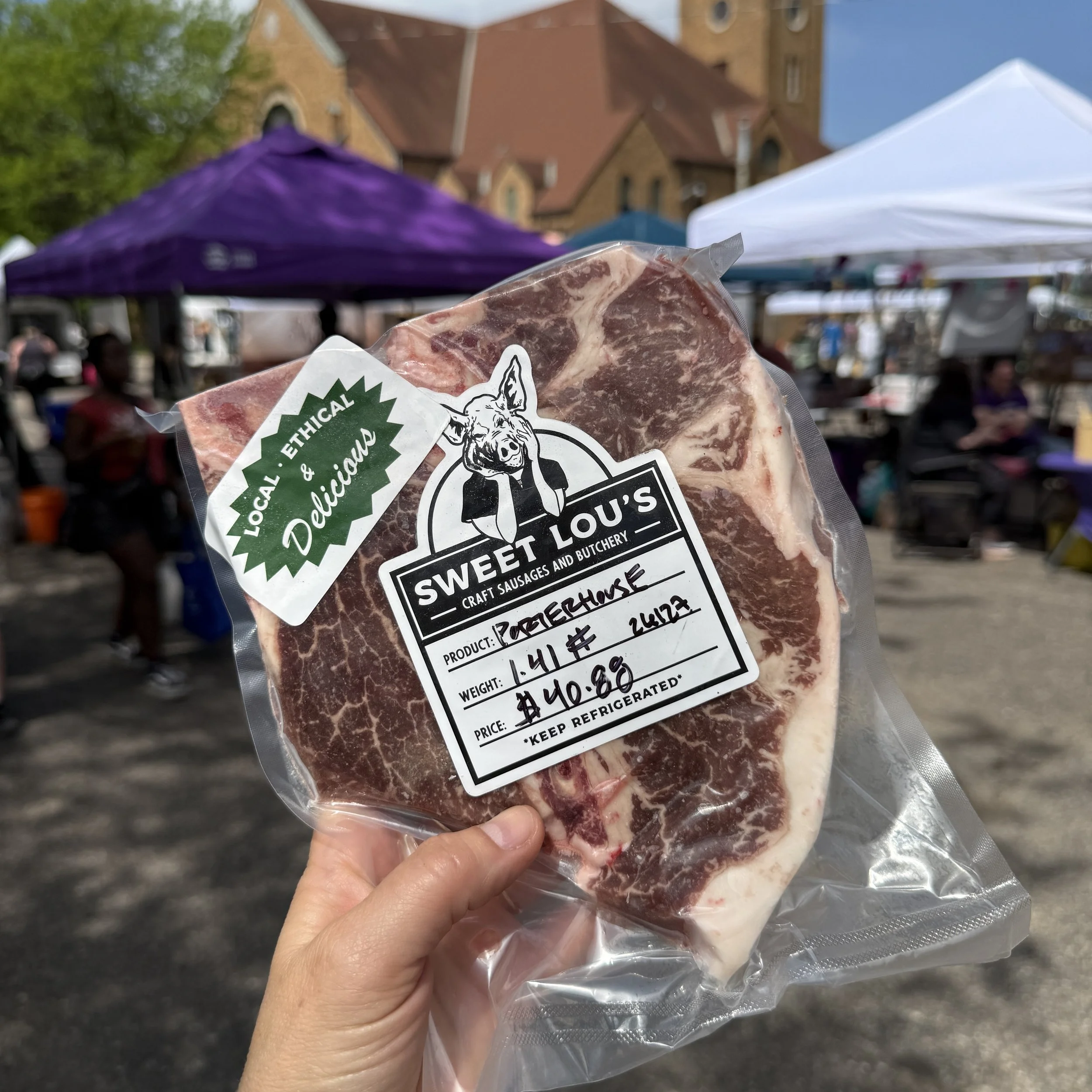A person holding a vacuum-sealed package of raw meat at an outdoor market with tents and a brick building in the background.