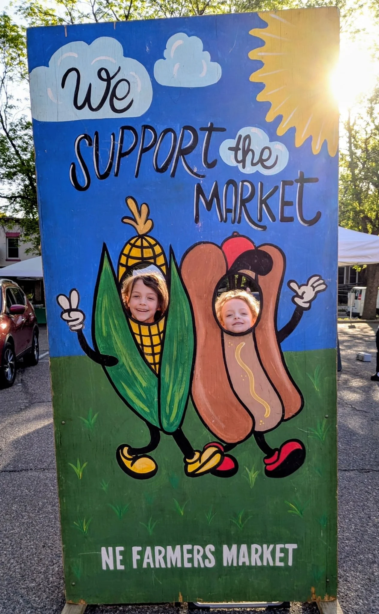 A colorful wooden sign at a farmers market features cartoon characters of a smiling corn cob and hot dog walking together, with children’s faces visible through cutouts in the sign. The sign promotes supporting the market and mentions local farmers.