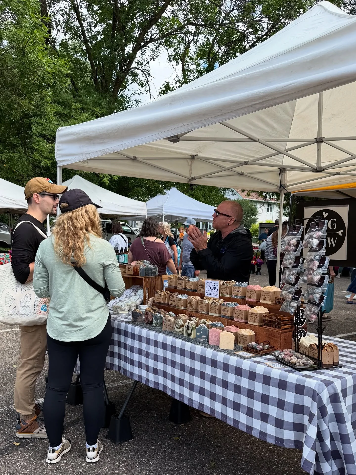 People browsing a soap and skincare product booth at an outdoor market, with trees and other vendor tents in the background.