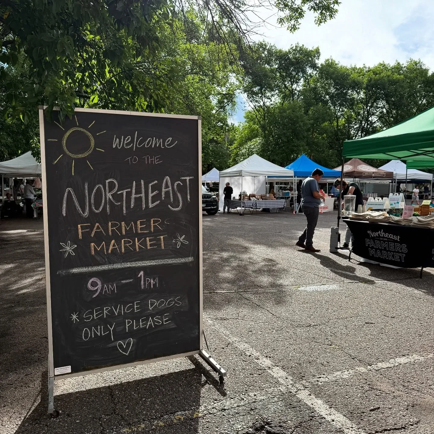 A chalkboard sign at the entrance of the Northeast Farmers Market, advertising the market hours from 9 AM to 1 PM and mentioning service dogs only. The market has multiple tents and vendors selling goods, with trees overhead and people shopping.