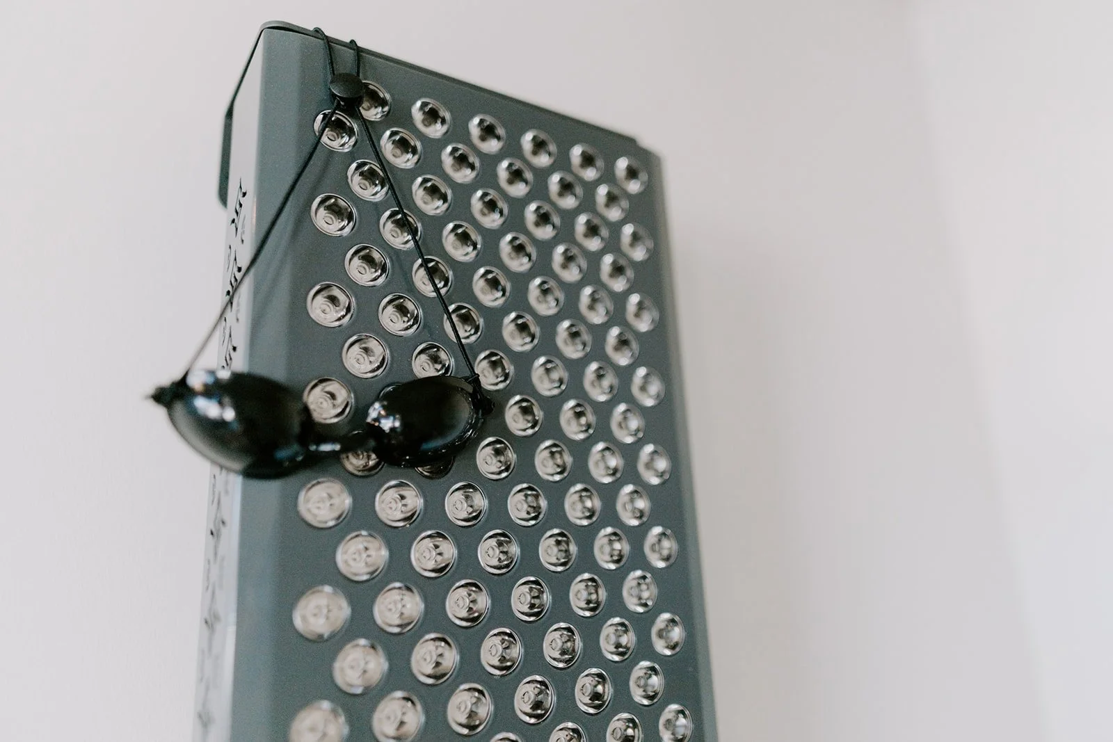 A pair of protective eye wear hanging on a red light therapy device, mounted on a white wall.