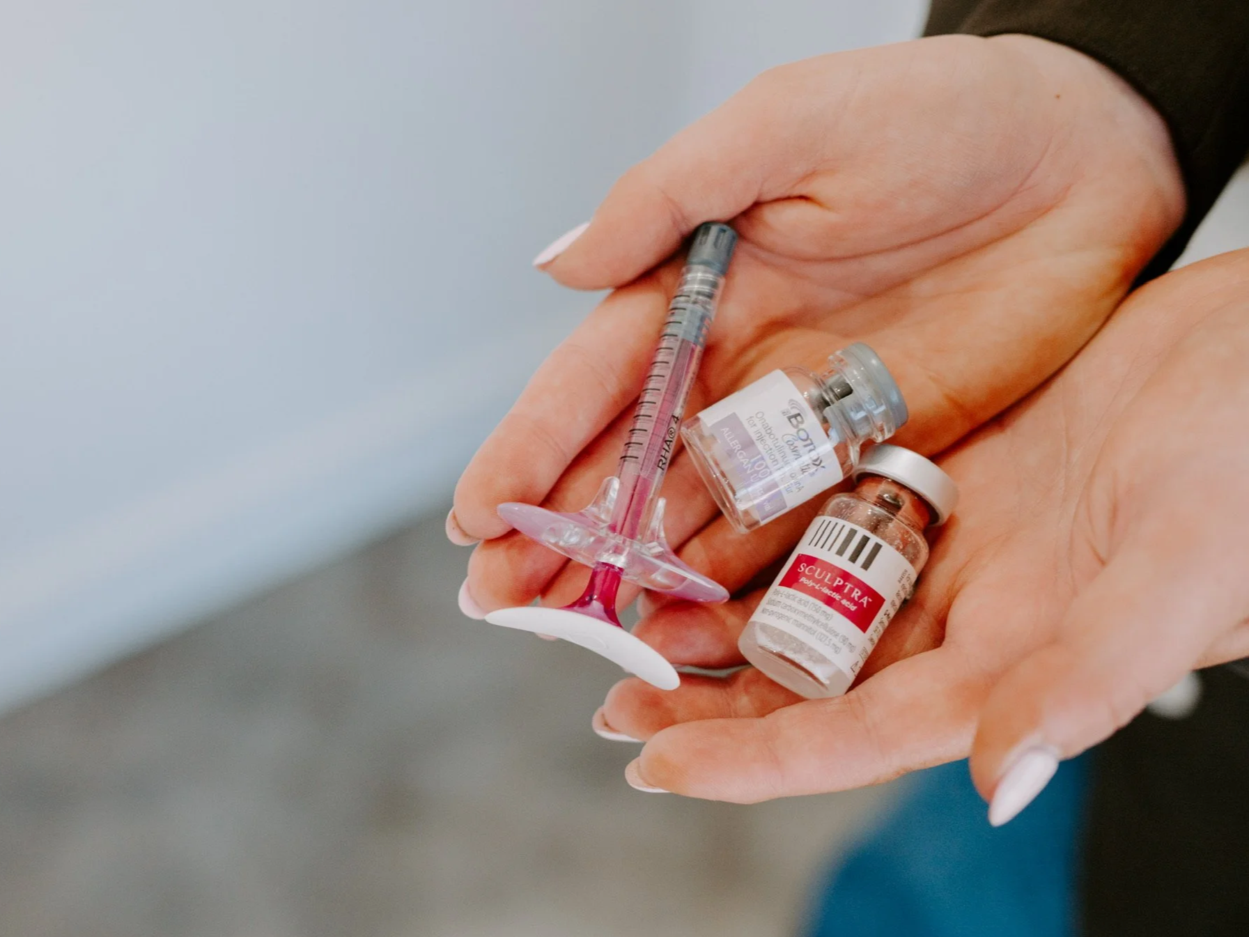 Hands holding a syringe, a small vial labeled 'Sculptra', and two other small vials, suggesting a medical or cosmetic procedure.