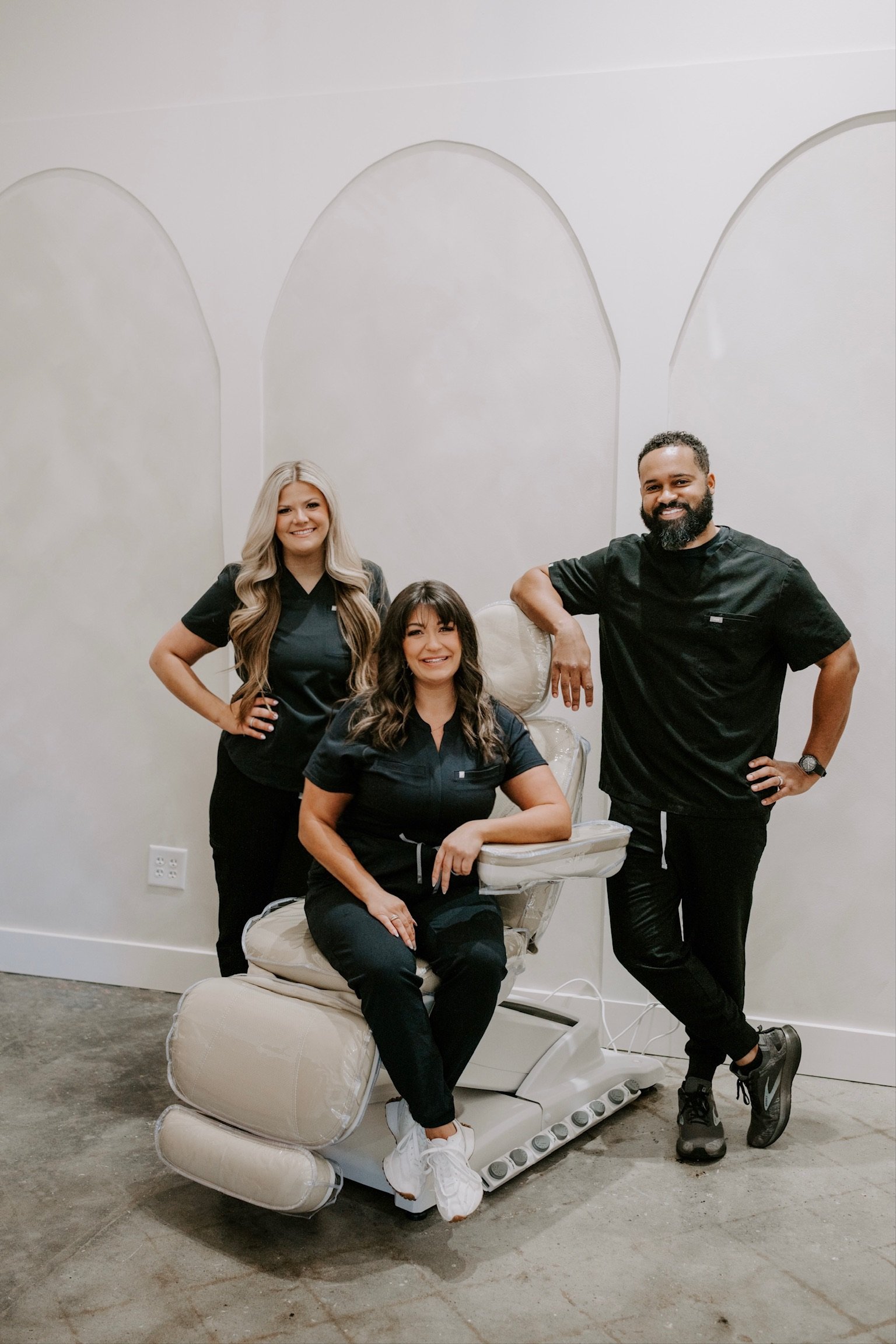 Three dental professionals, two women and one man, in black scrubs posing with a dental chair in a clean, modern office.