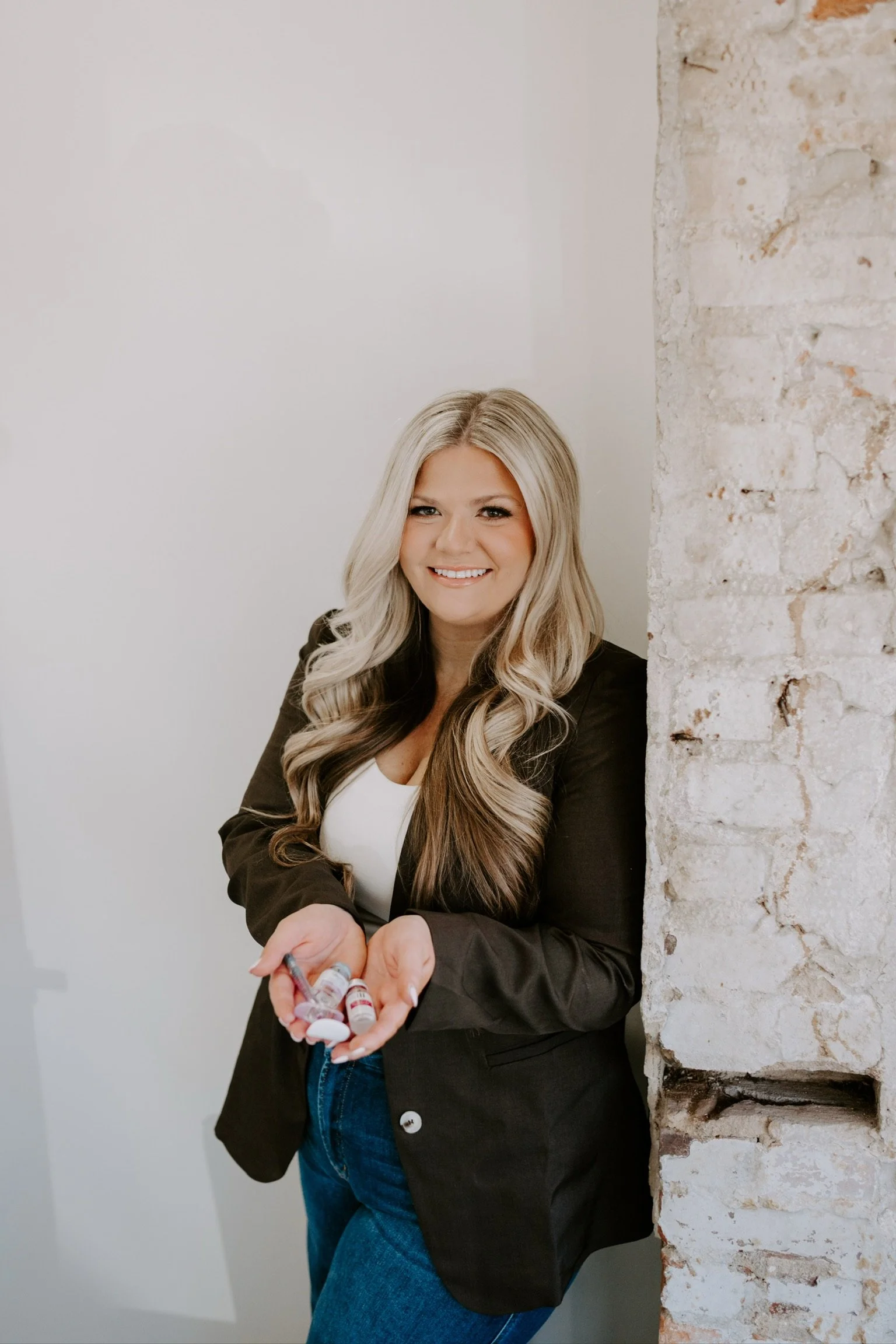 A woman with long blonde hair, smiling, holding prescription pill bottles in her hand, leaning against a rustic brick wall.
