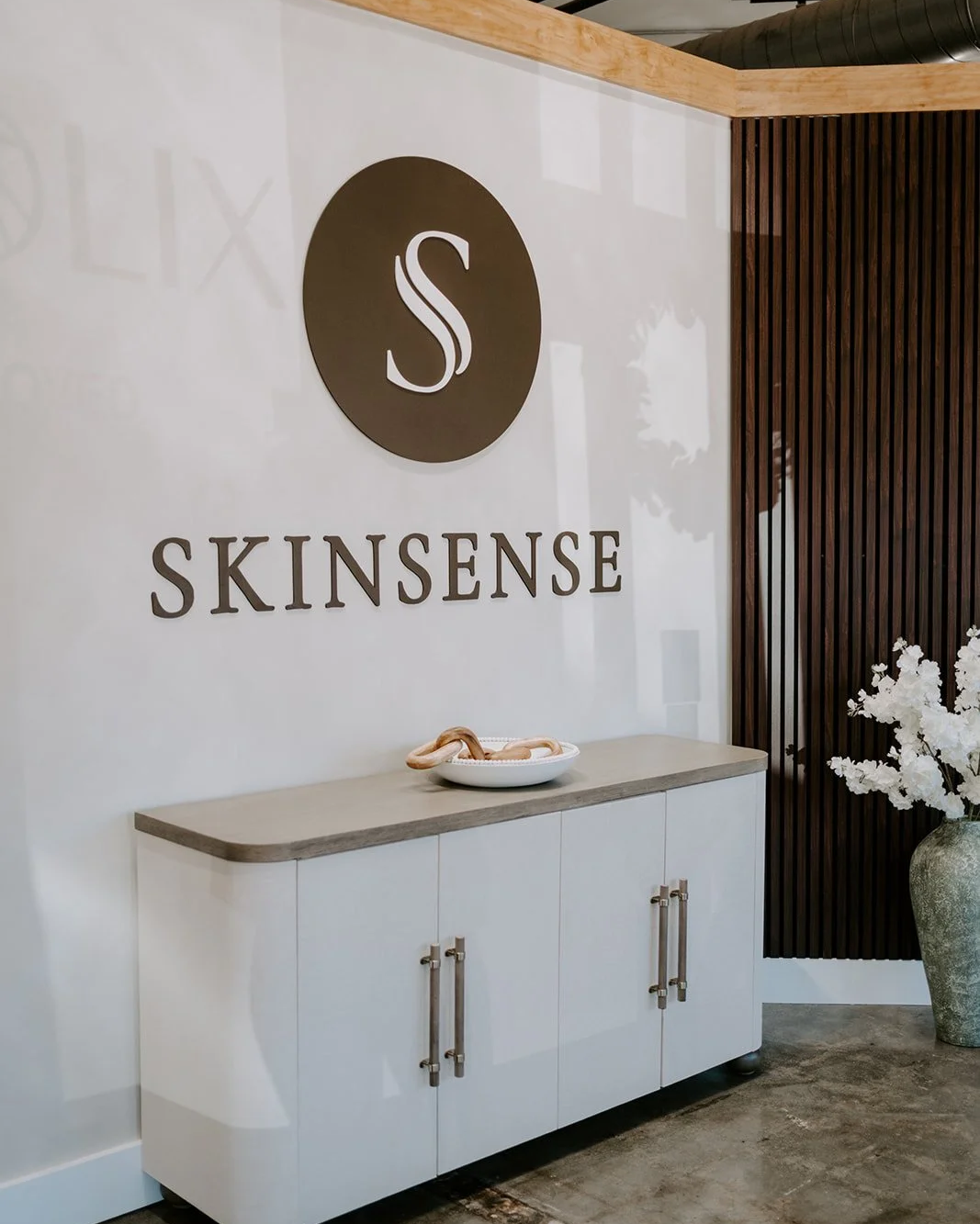 Interior of Skinsense spa or skincare clinic with a white cabinet, a decorative snake on a tray, and a large vase with white flowers next to a dark wooden wall panel.