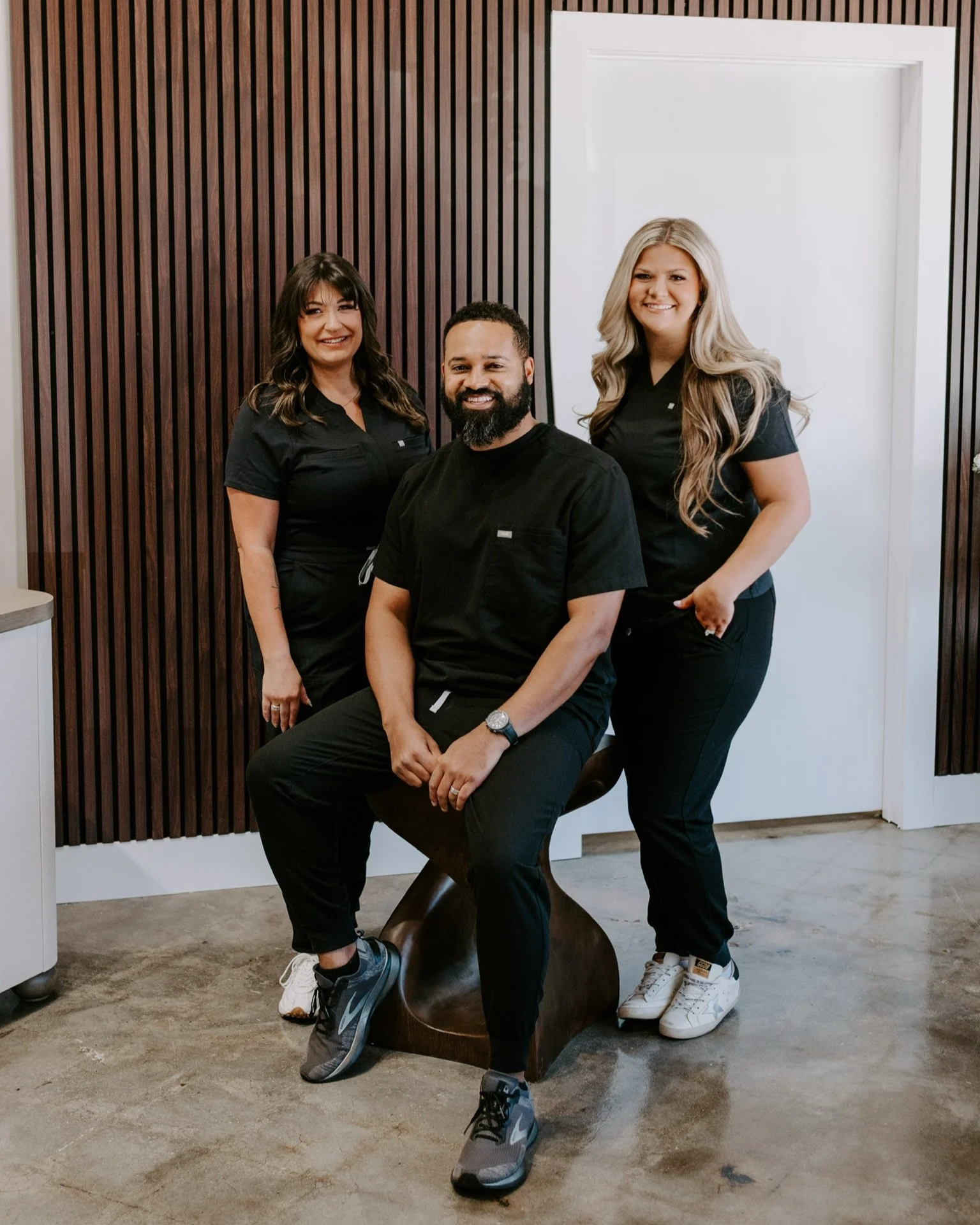 Three people, two women and one man, wearing black scrubs in a room with wooden wall paneling and a white door, smiling for the photo. The man is sitting on a modern wooden stool, while the women stand on either side.