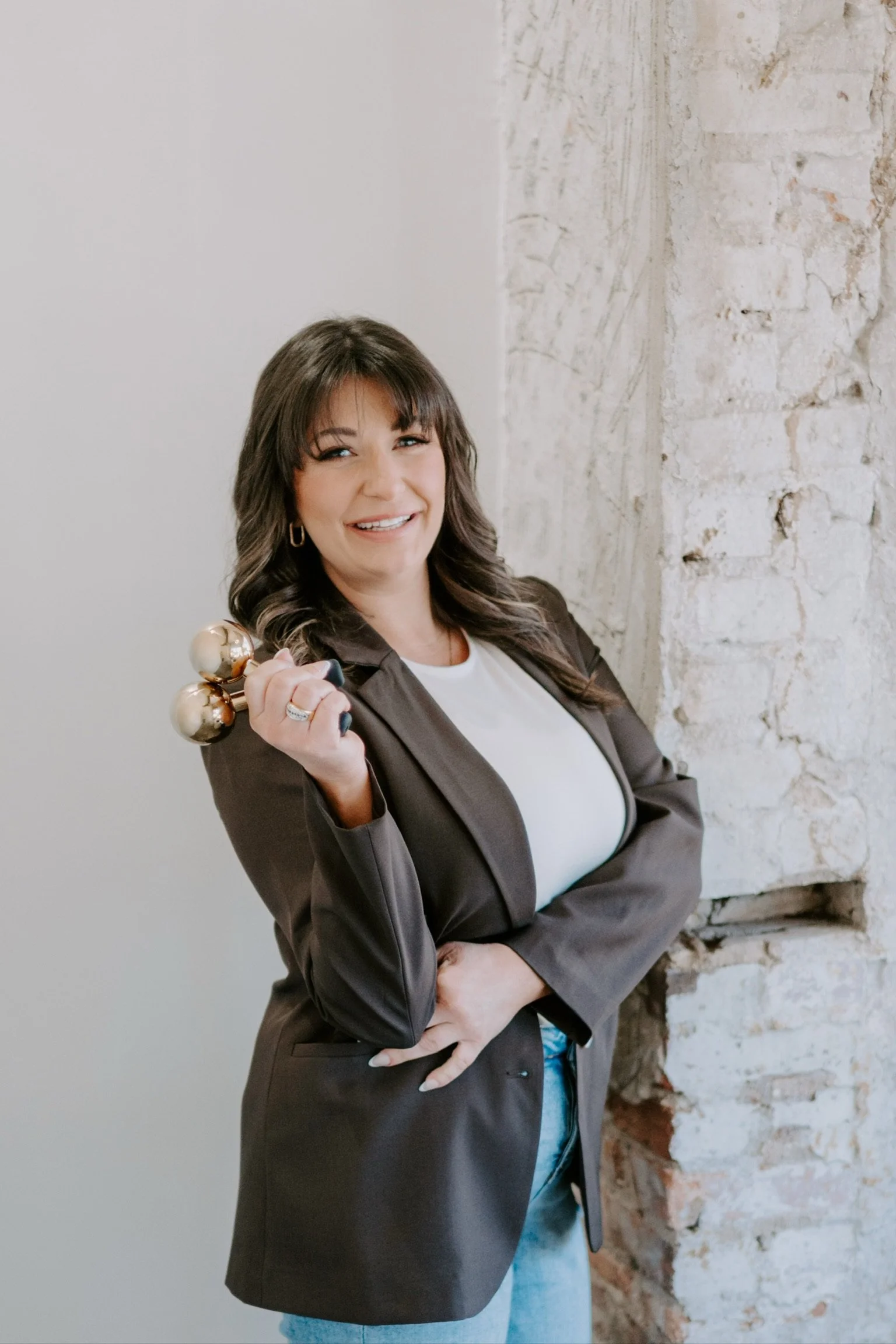 A woman with dark brown hair, wearing a brown blazer over a white top, smiling and holding a decorative gold and pearl lamp in her right hand, standing against a white brick wall.