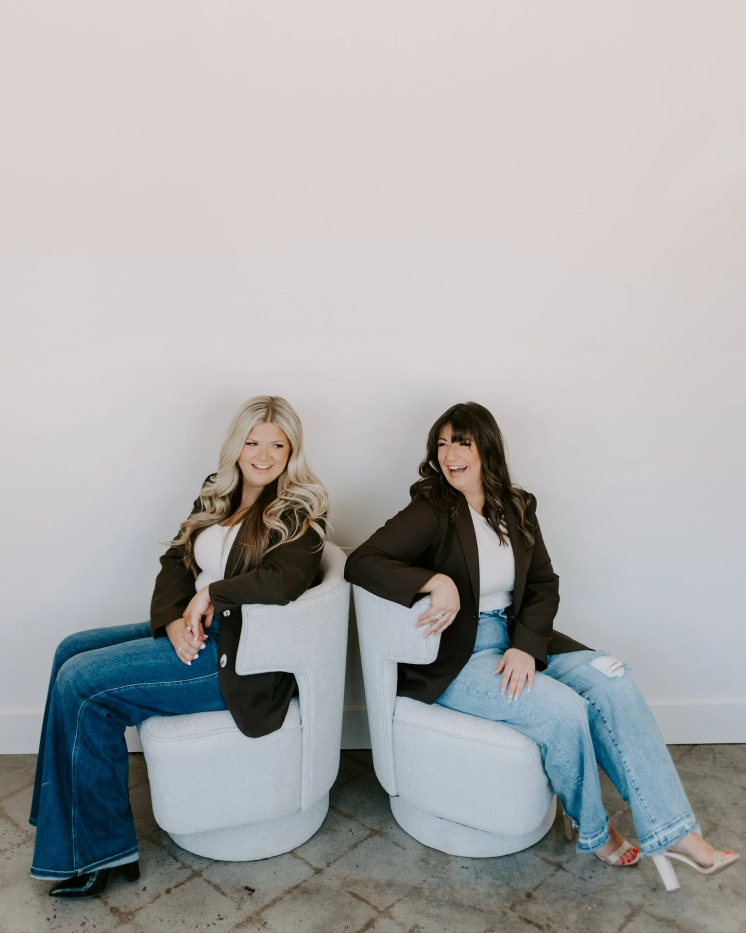 Two women sitting back to back on white chairs, smiling and laughing, against a white wall.