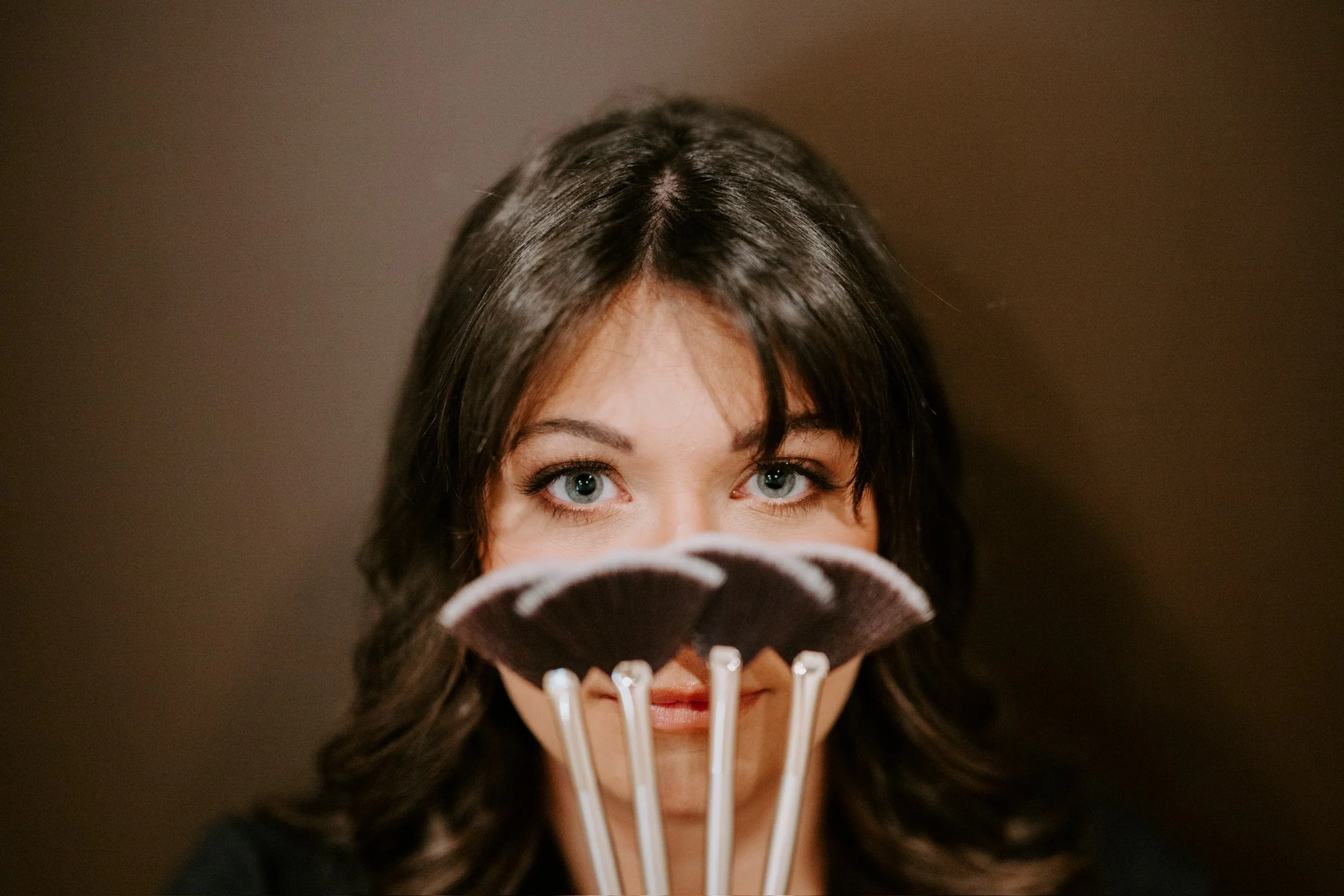 Woman with dark hair and blue eyes holding a fan of makeup brushes in front of her face, partially covering her face.