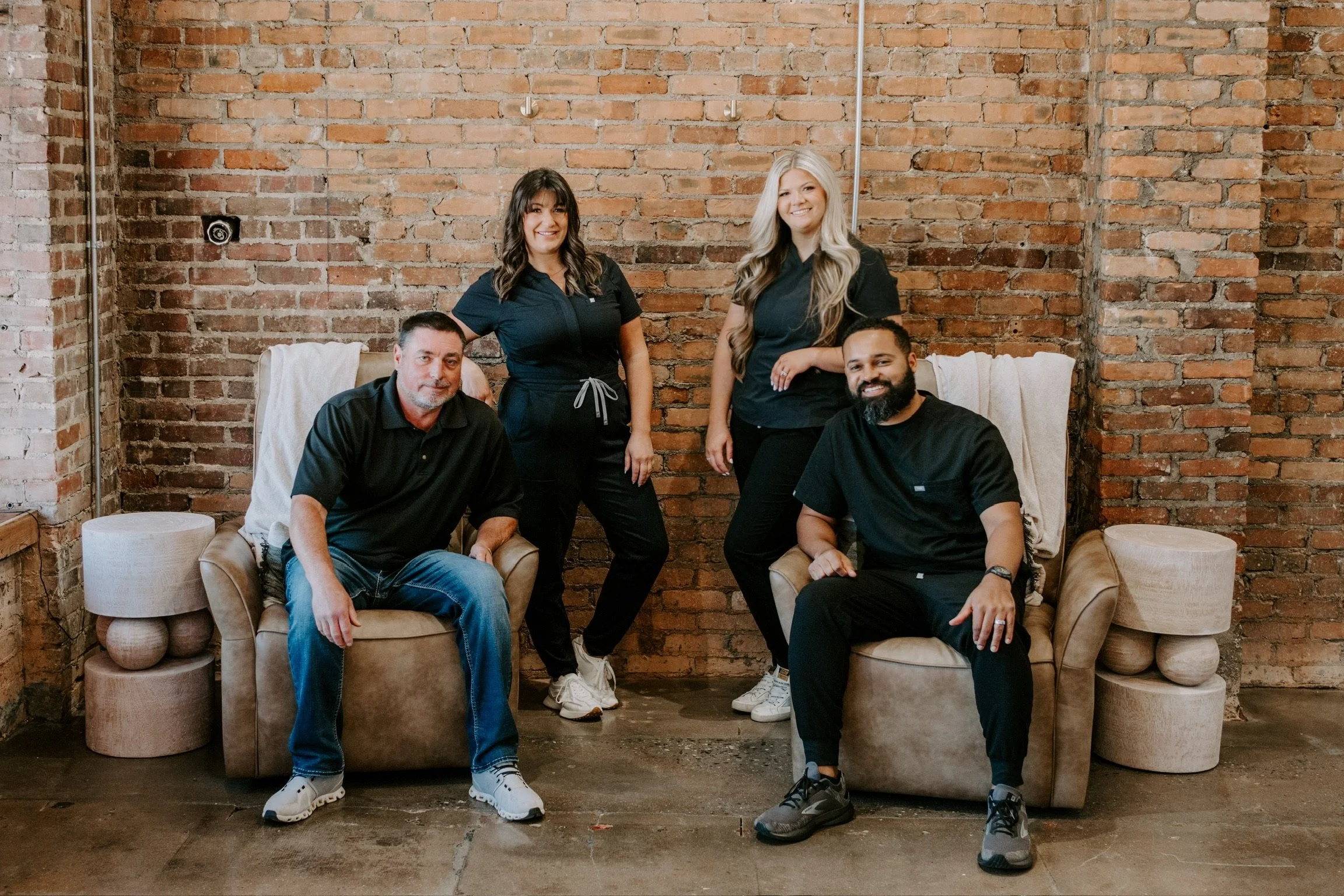 Group of four people in black clothing sitting and standing in front of a brick wall in a studio setting