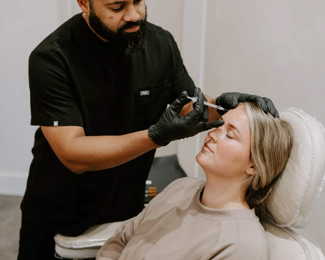 A woman is lying on a treatment chair with her eyes closed while a healthcare professional, wearing black gloves, administers a cosmetic injection to her forehead.
