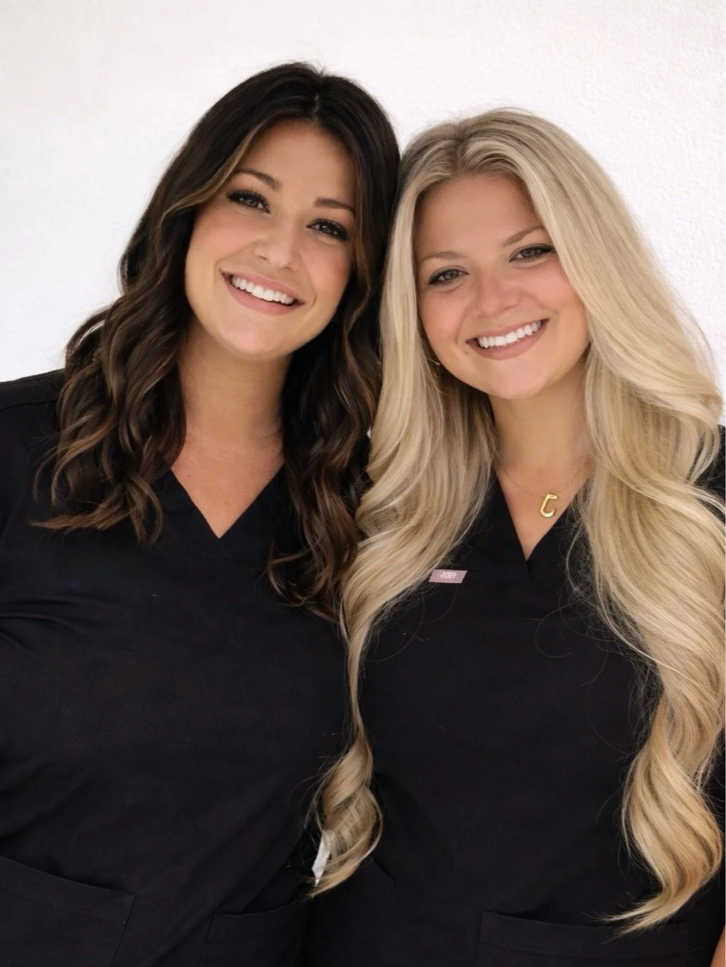 Two women with long hair smiling, standing close together, wearing black medical scrubs, against a plain white wall.