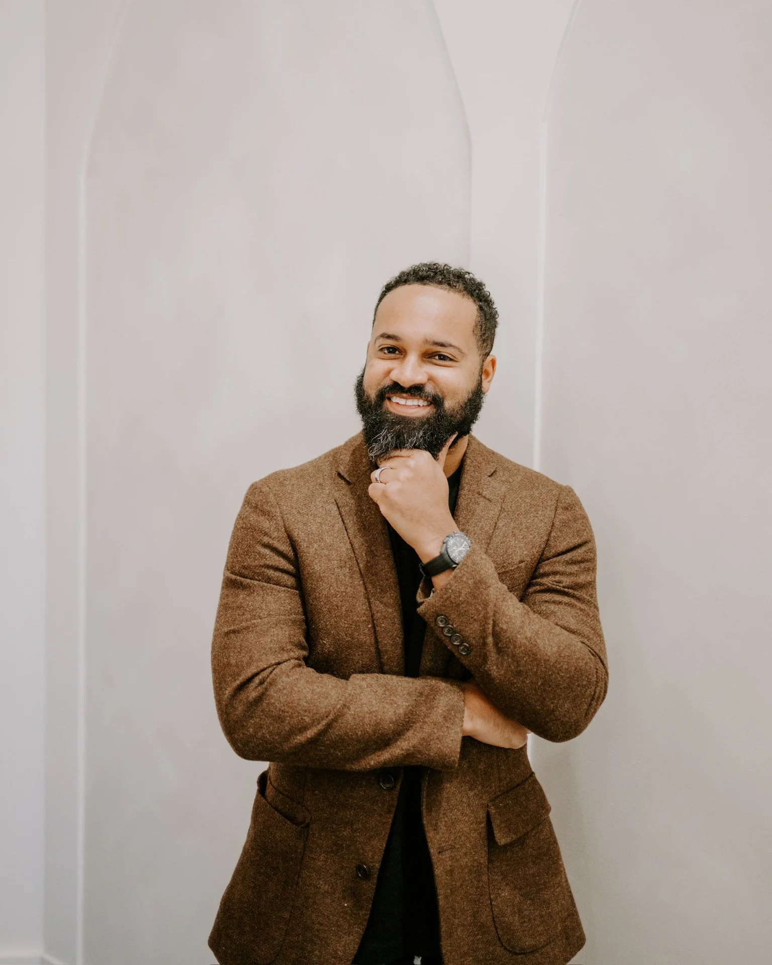 A man with a beard and short curly hair, dressed in a brown blazer and black shirt, smiling and posing with his hand on his chin, standing against a plain light-colored wall.