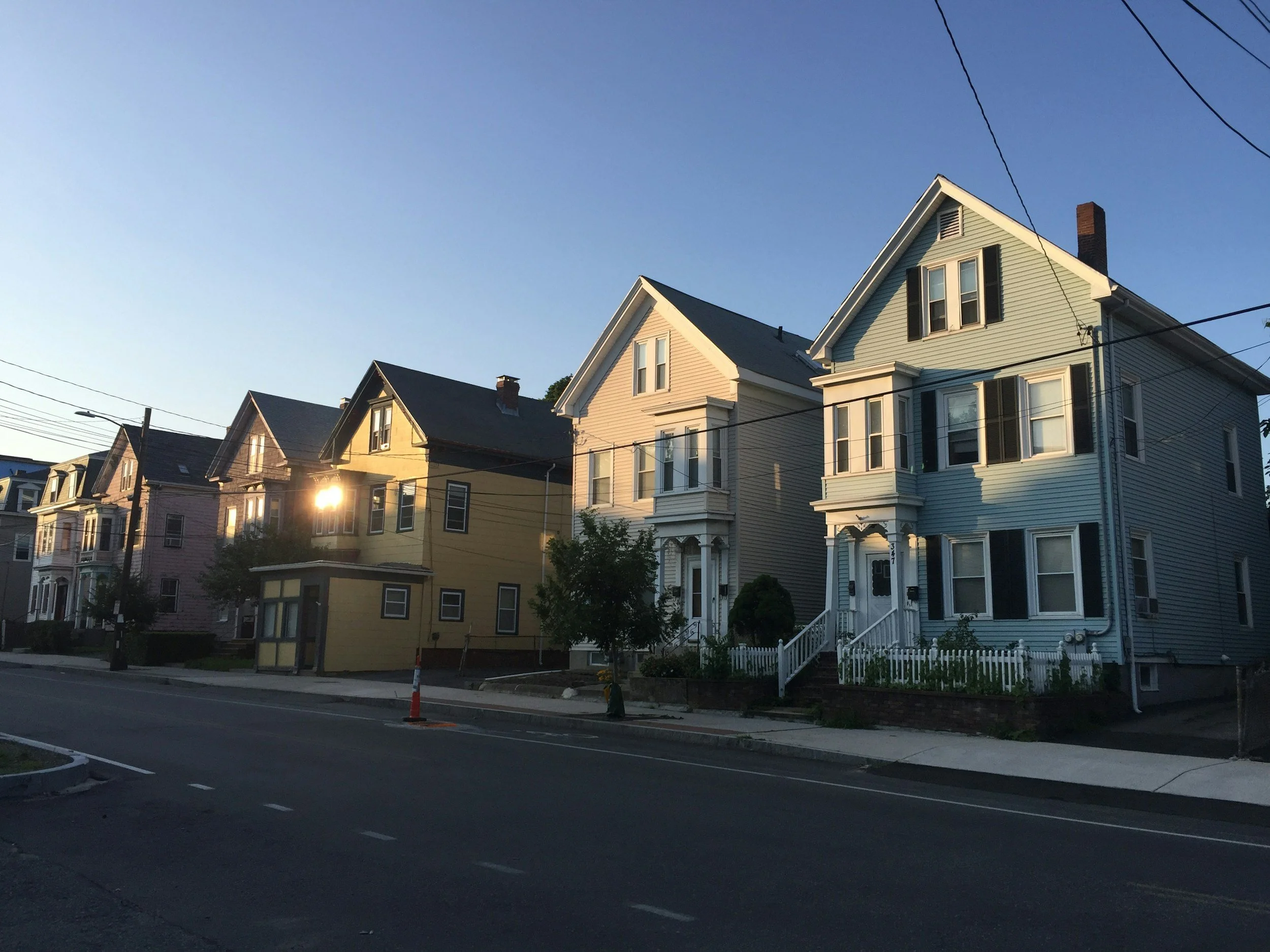 A row of colorful houses on a street during dusk with clear sky and streetlights.