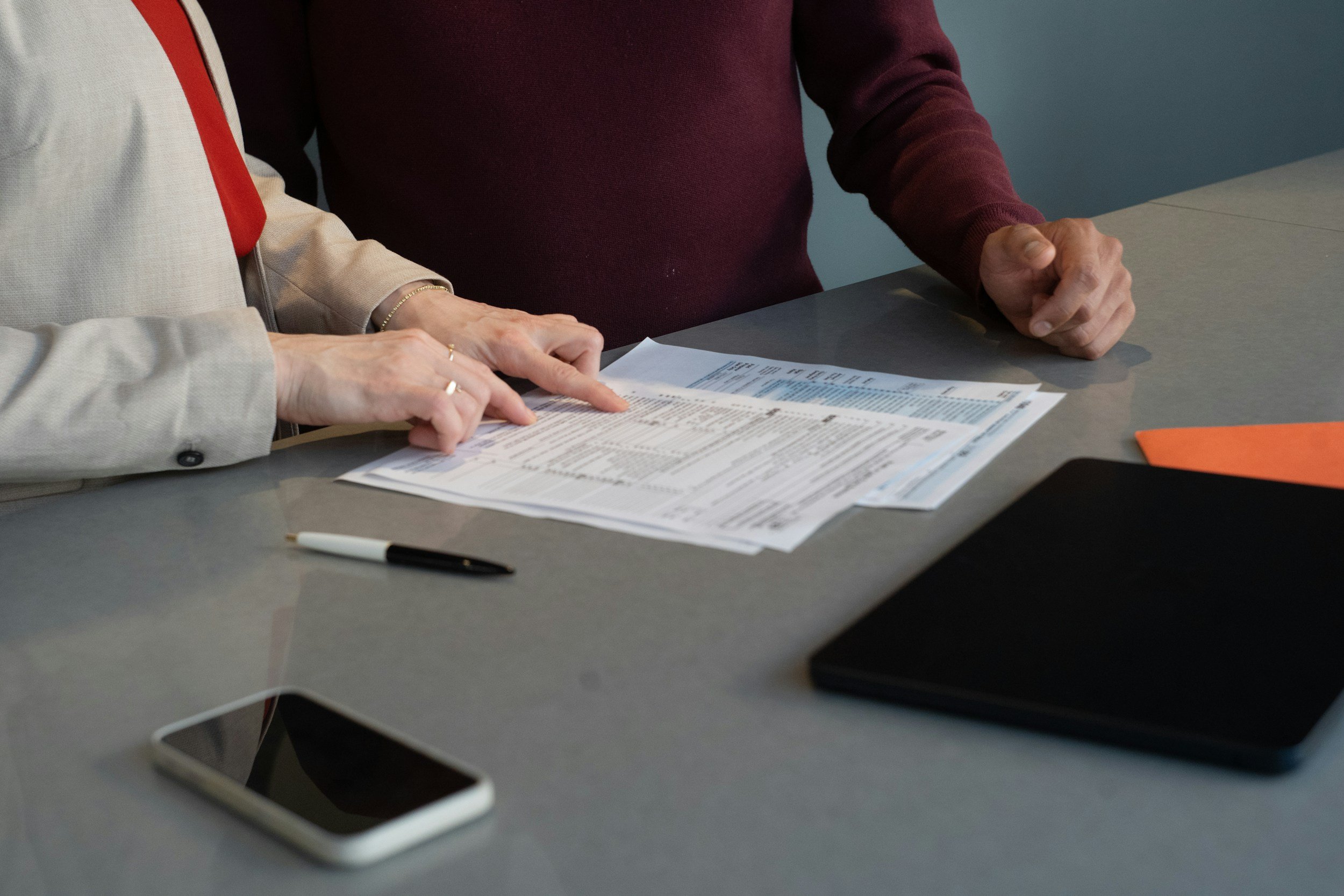 Two people sitting at a table, discussing documents. Only their hands and torsos are visible. One person is pointing at the papers.