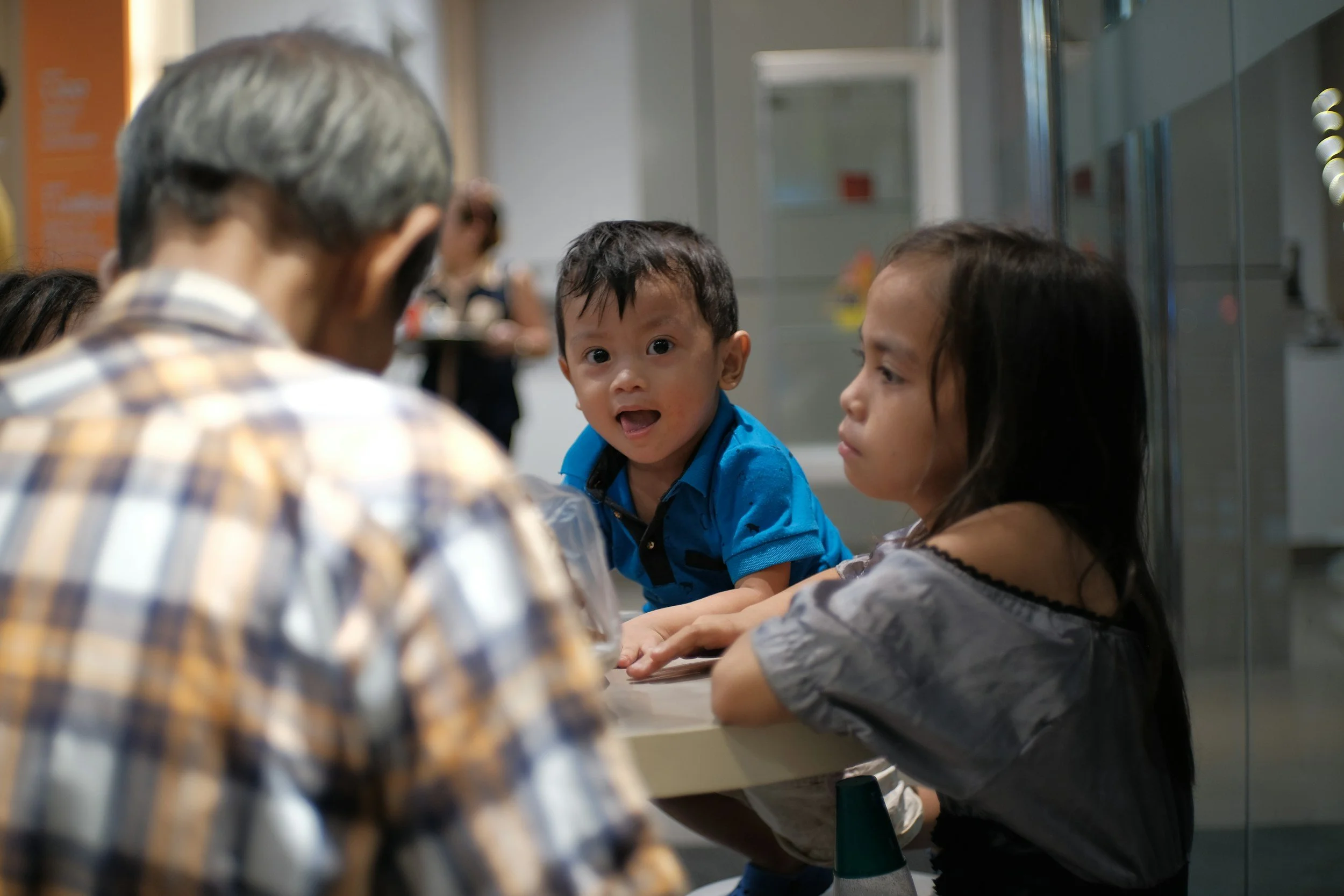 Four children sitting at a table in a school hallway, one boy in a blue shirt looking at the camera and an older woman with gray hair and a plaid shirt focused on a task. Lockers are in the background.