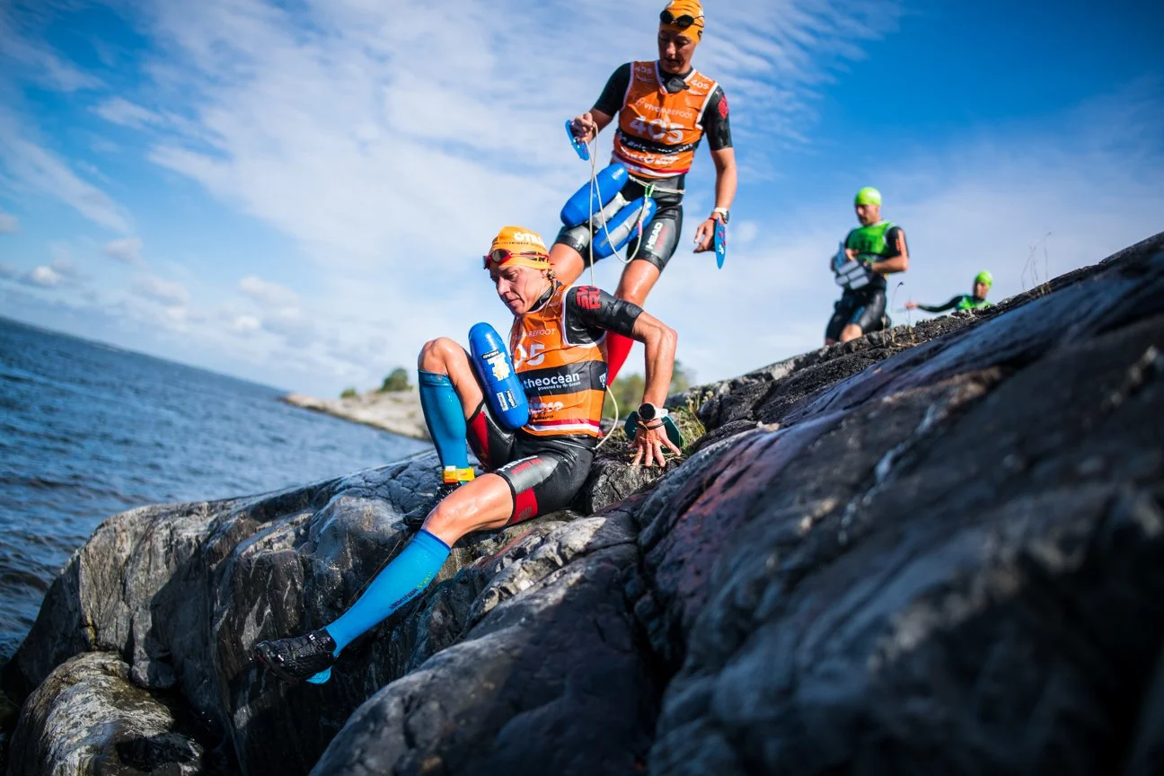 Triathlon athletes climbing over rocks near a body of water, with one athlete helping another while others are in the background; all are wearing wetsuits, swim caps, and carrying gear.