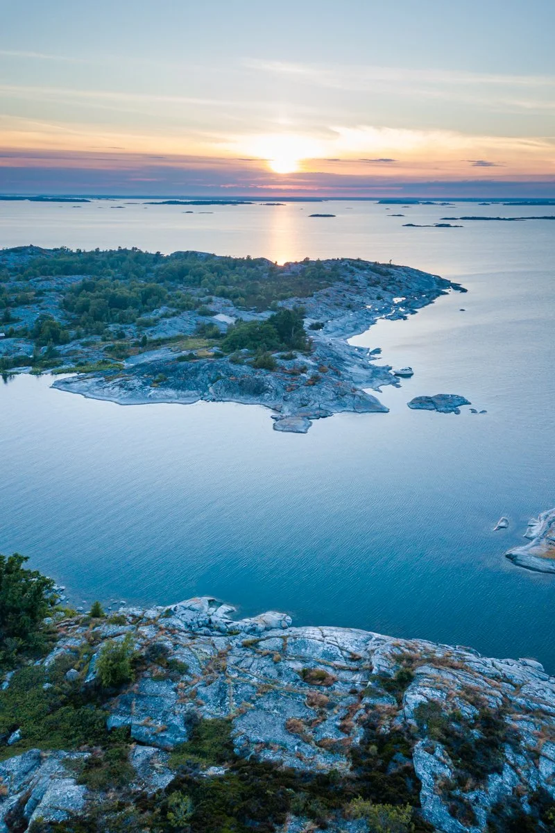Aerial view of a rocky island with trees, surrounded by a calm body of water, during sunset with the sun partially visible on the horizon.