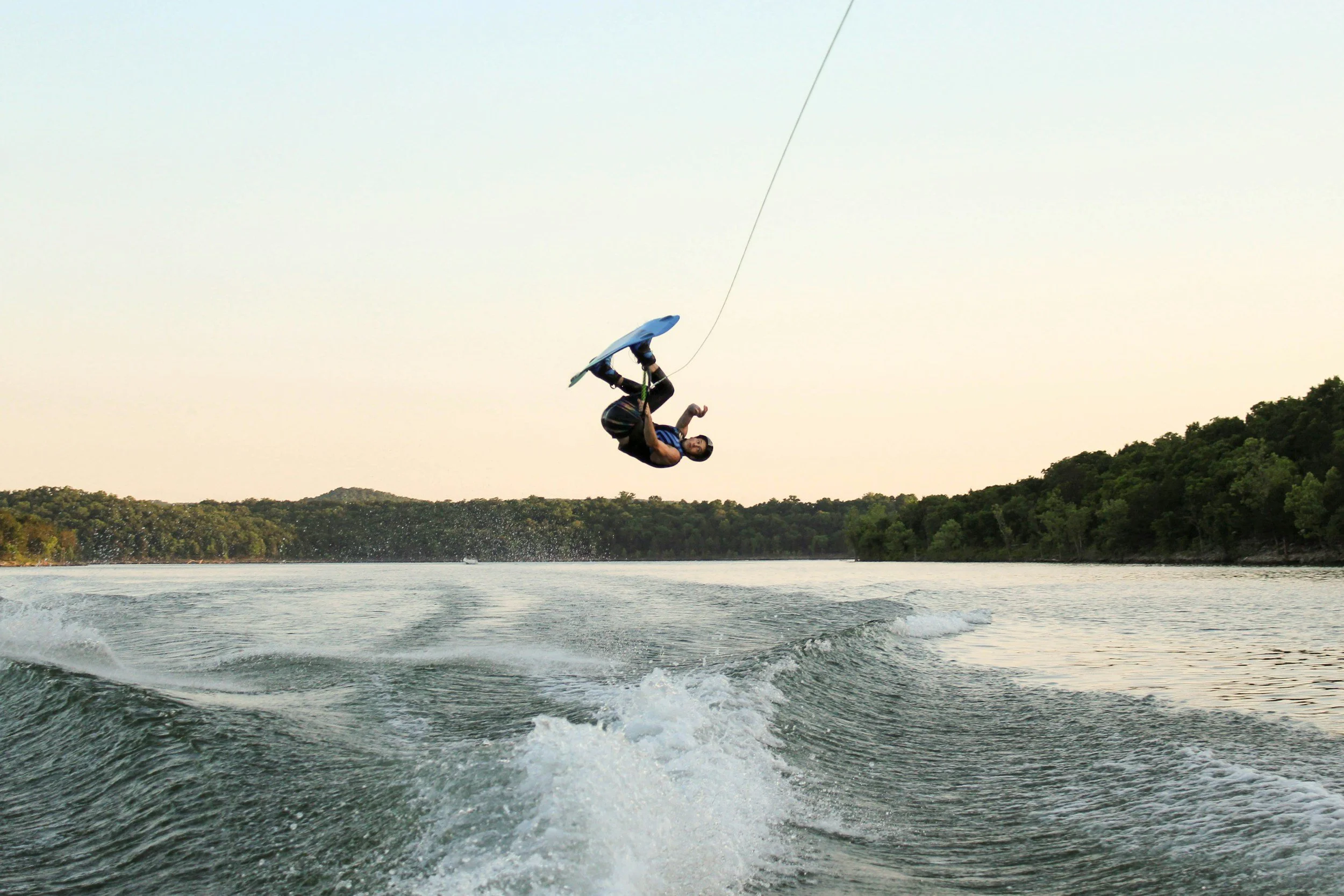 A person performing a flip while wakeboarding on a lake, with trees in the background and a clear sky.