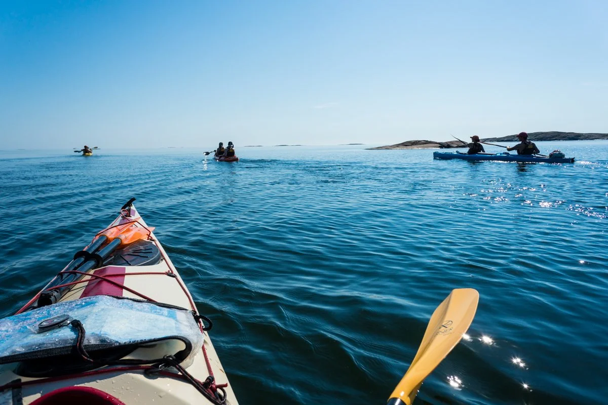 View from a kayak with a paddle, looking out at two other kayakers paddling near an island and a boat in the distance on a sunny day.