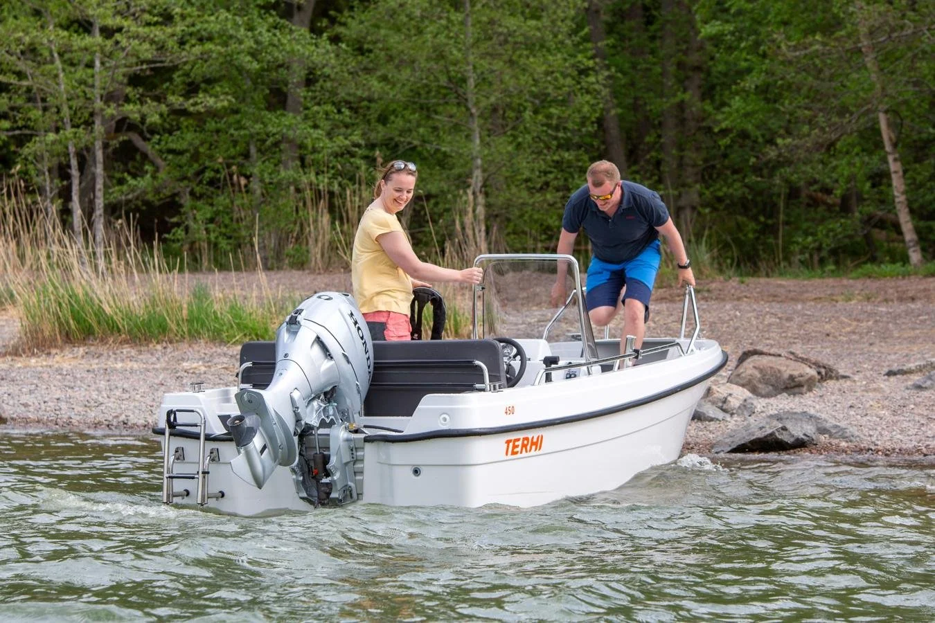 Two people, a woman and a man, on a small white motorboat near a rocky shoreline with green trees in the background, smiling and preparing to depart on the water.