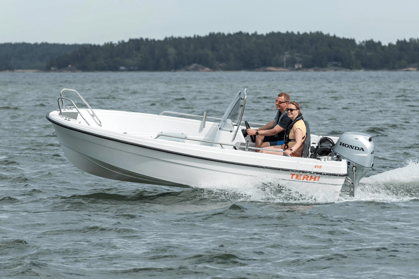 En vit motorbåt med två personer ombord, en man och en kvinna, som snabbt rör sig över ett vattendrag med en skogsklädd strandlinje i bakgrunden.