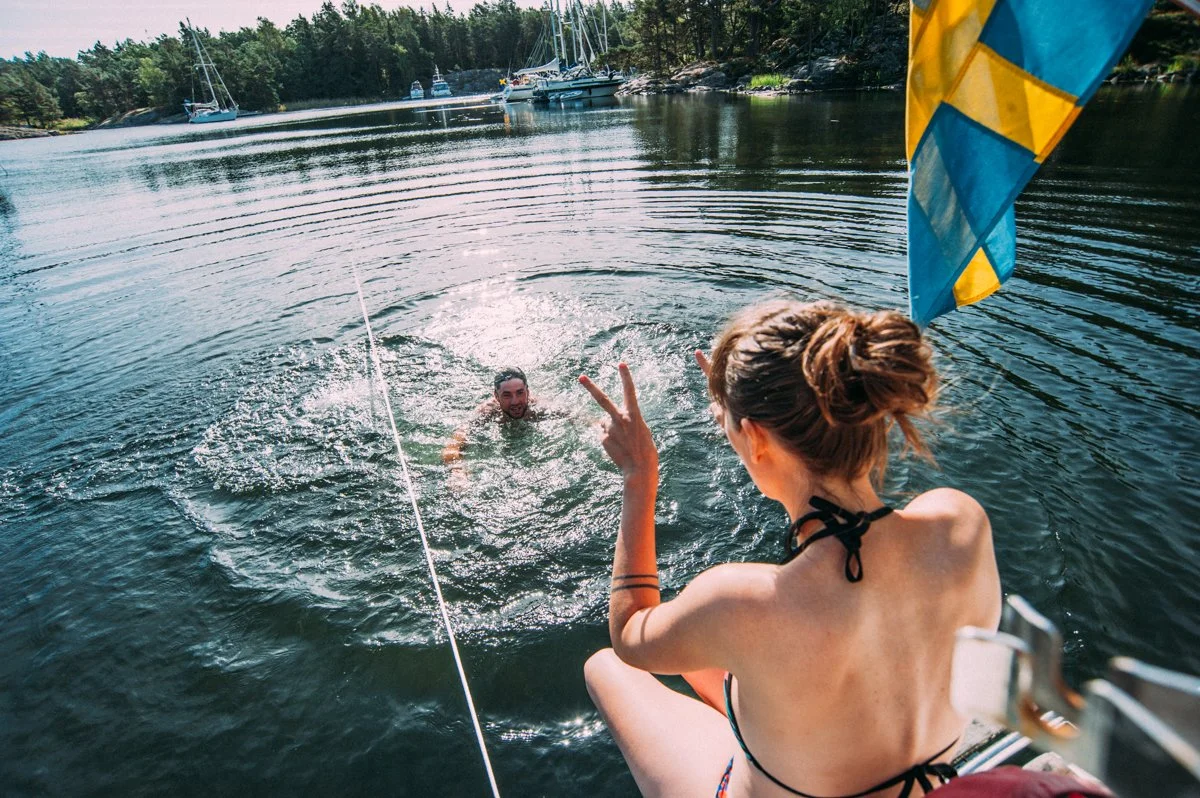 Two people on a boat. One person with braided hair making a peace sign, the other swimming in the water near the boat. Several sailboats and trees on a shoreline in the background.