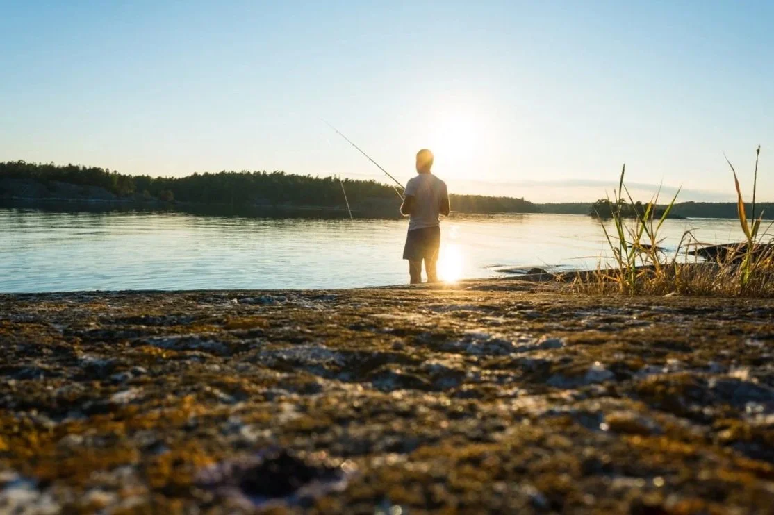 A child fishing by a lake at sunset, standing on the rocky shore with trees in the background.
