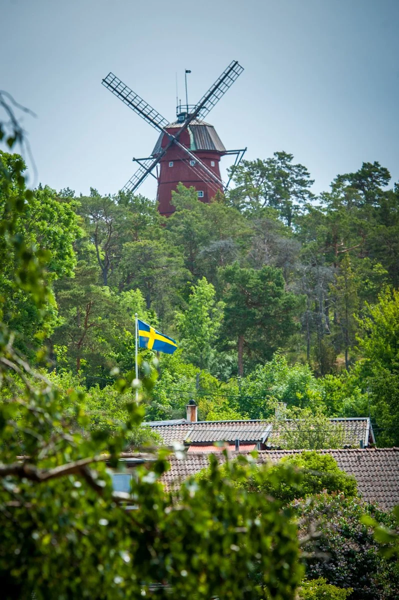 A red windmill on a hill surrounded by green trees, with a Swedish flag flying in a residential area below.