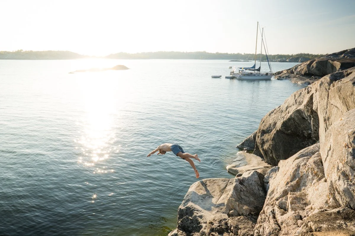 A person jumping off rocks into a calm body of water, with sailboats anchored nearby and a bright sun reflecting off the water.