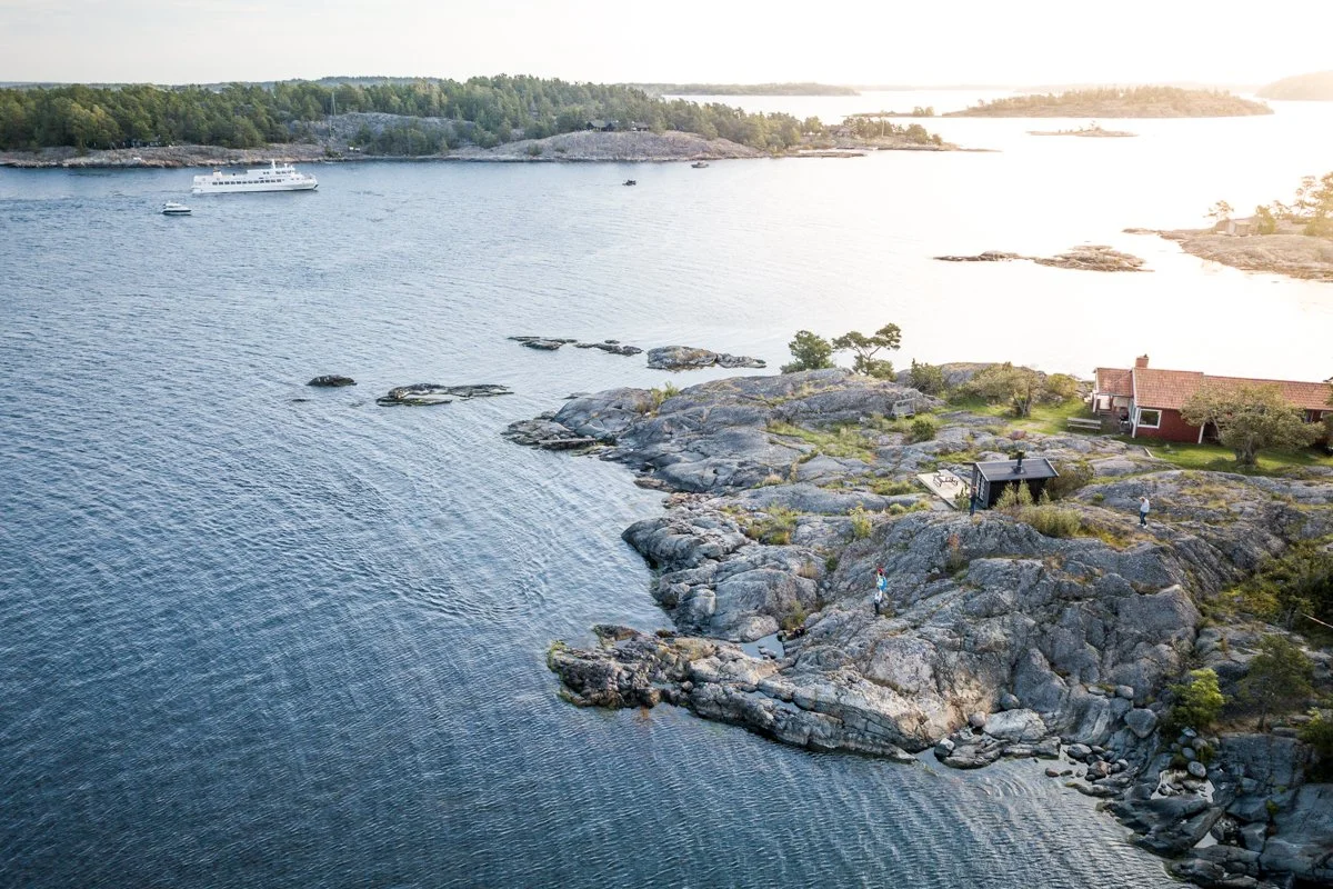 A scenic coastal view with rocky shoreline, calm water, a few people exploring, a red house, and a boat docked in the distance on a sunny day.
