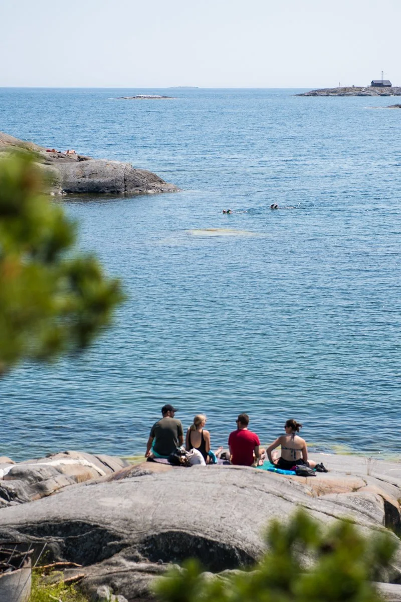 Four people sitting on rocks by a body of water, with two other people swimming in the water nearby. The water is calm and blue, with small islands and a lighthouse in the distance.