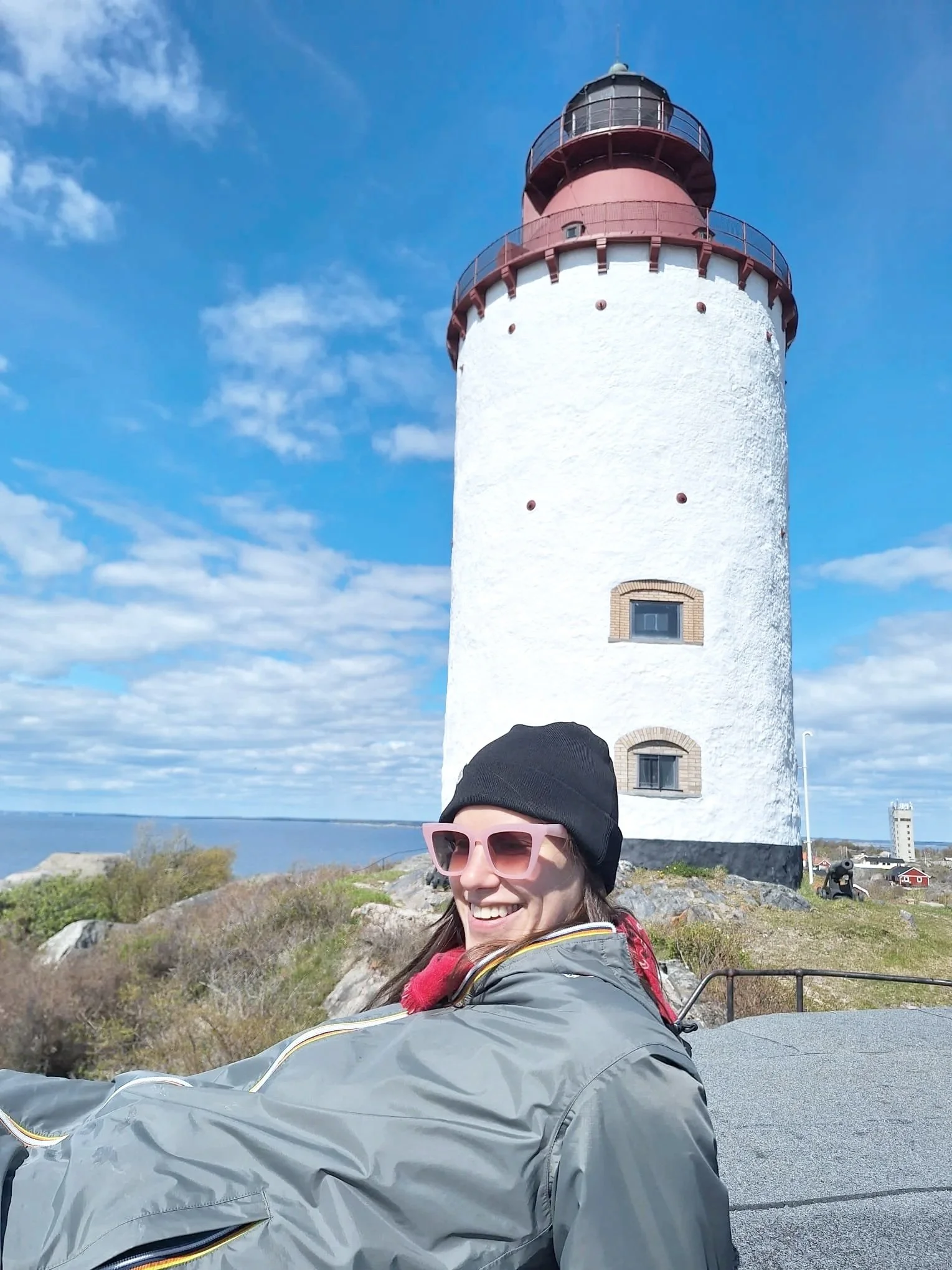A woman in a gray jacket, pink sunglasses, and a black beanie smiling in front of a tall white lighthouse with a red top, on a rocky landscape with a blue sky and clouds.