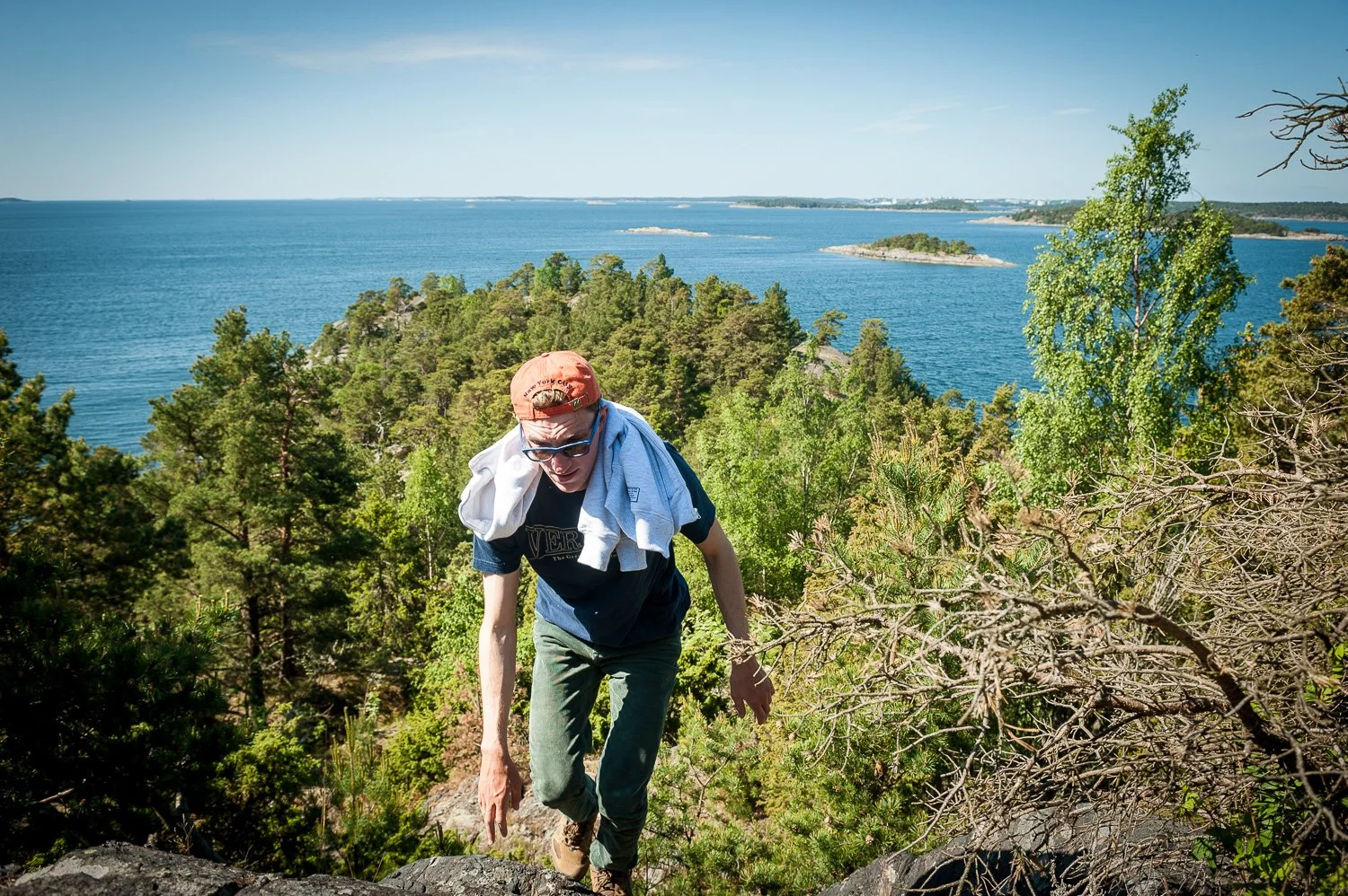 A person hiking up a rocky incline surrounded by trees, with a view of a large body of water with small islands in the distance.