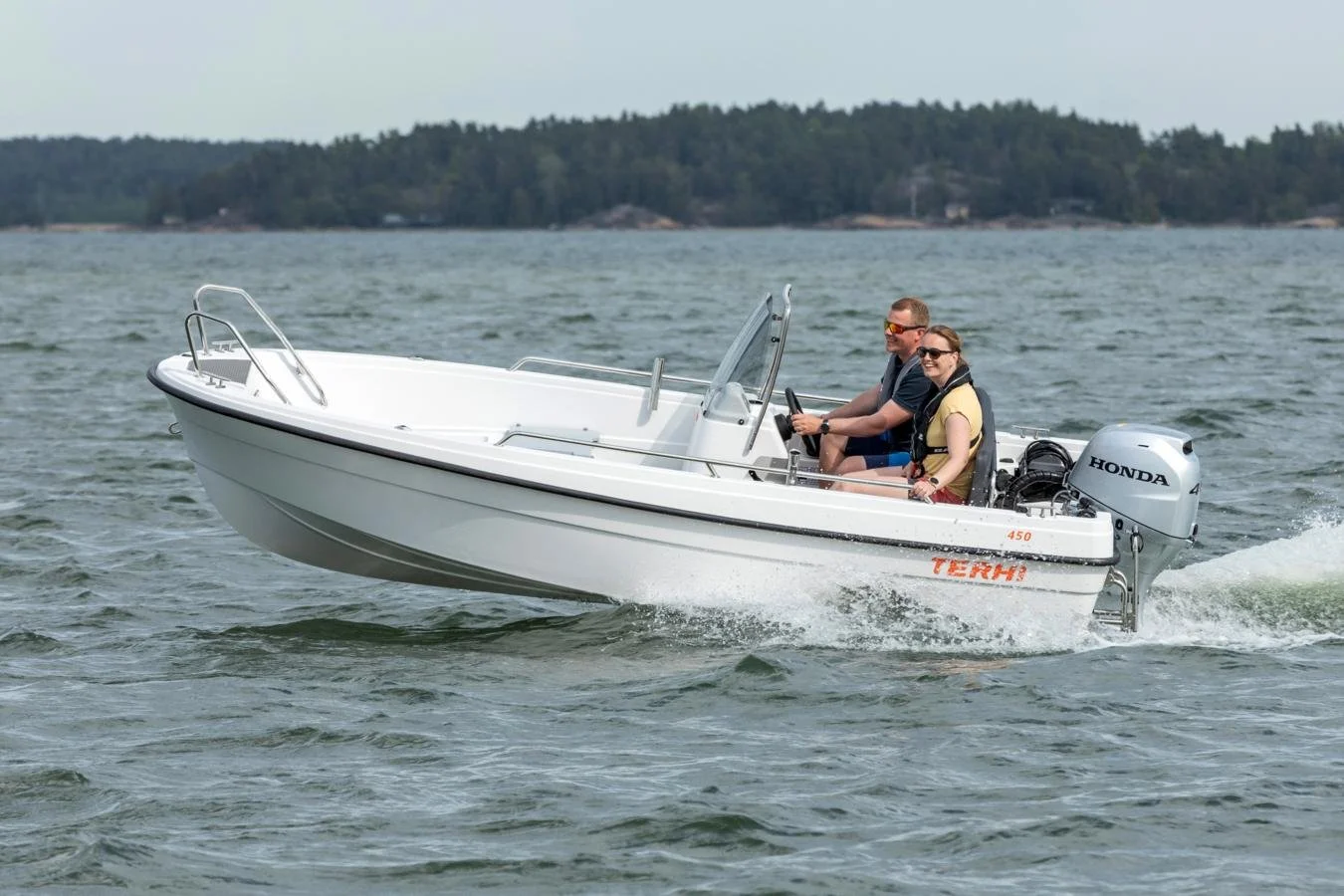 Two people in a small white motorboat on the water, with a forested shoreline in the background.