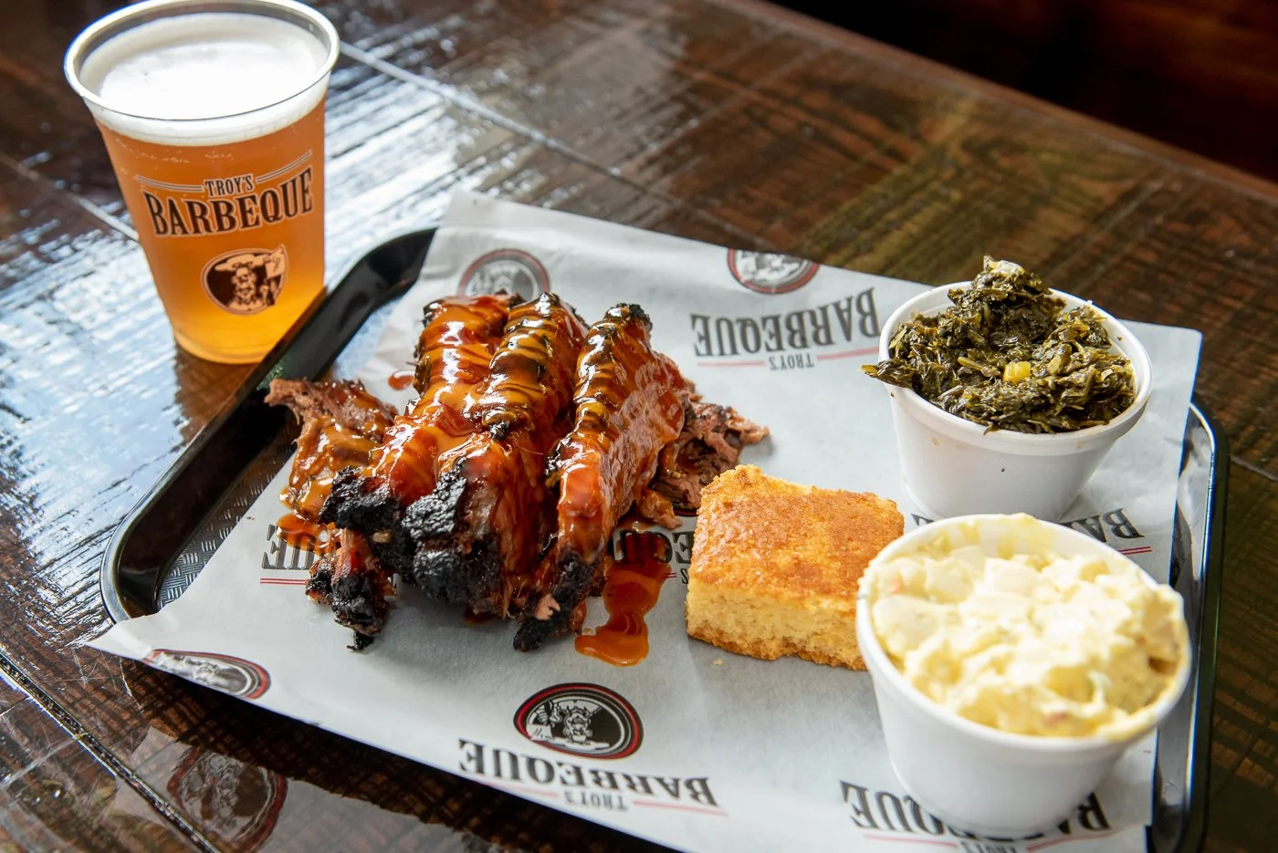 Barbecue meal on a tray with ribs, cornbread, collard greens, macaroni and cheese, and a glass of beer at Troy's Barbeque restaurant.