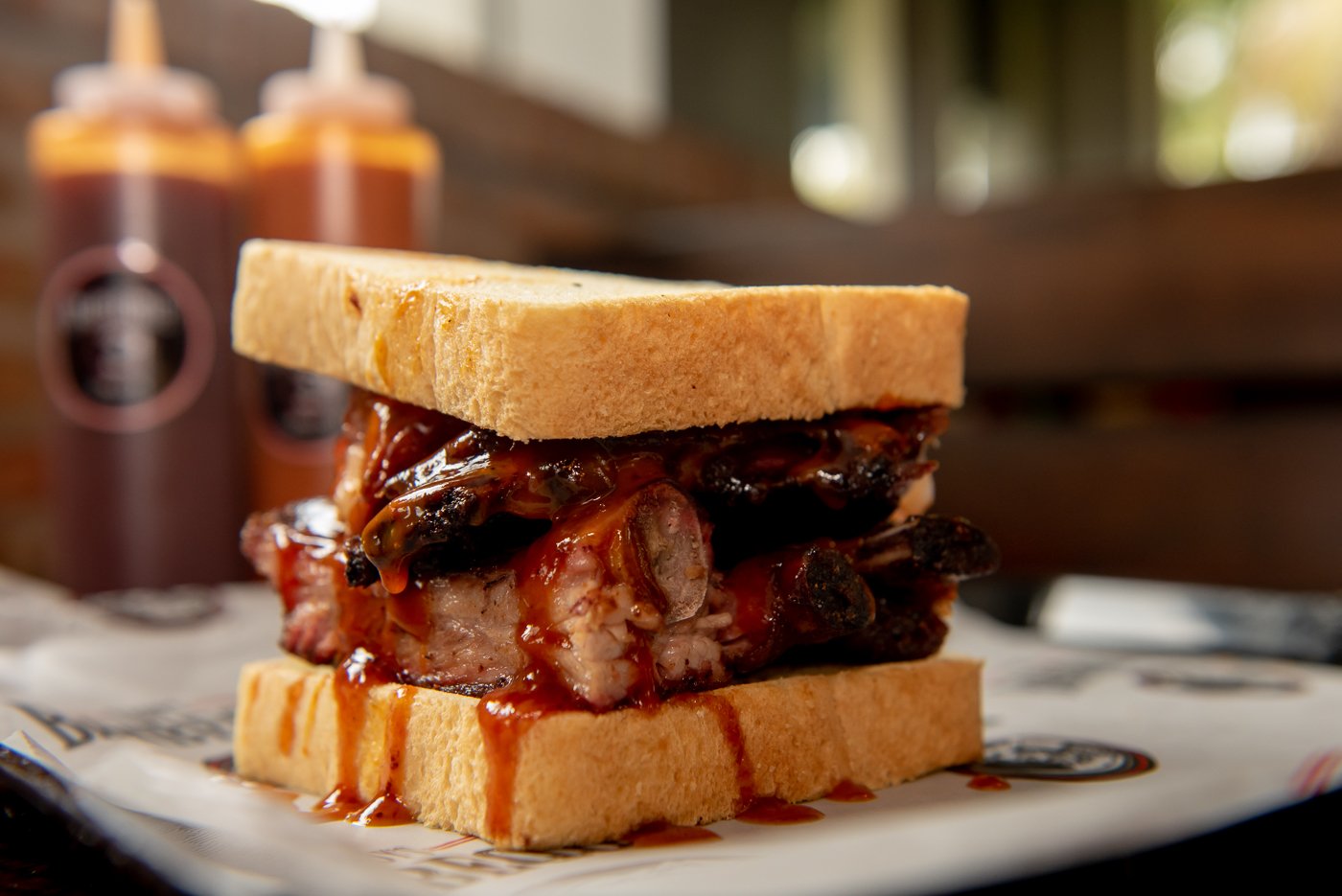 Barbecue sandwich with pork, barbecue sauce, and toasted bread slices on paper with bottles of barbecue sauce in the background.
