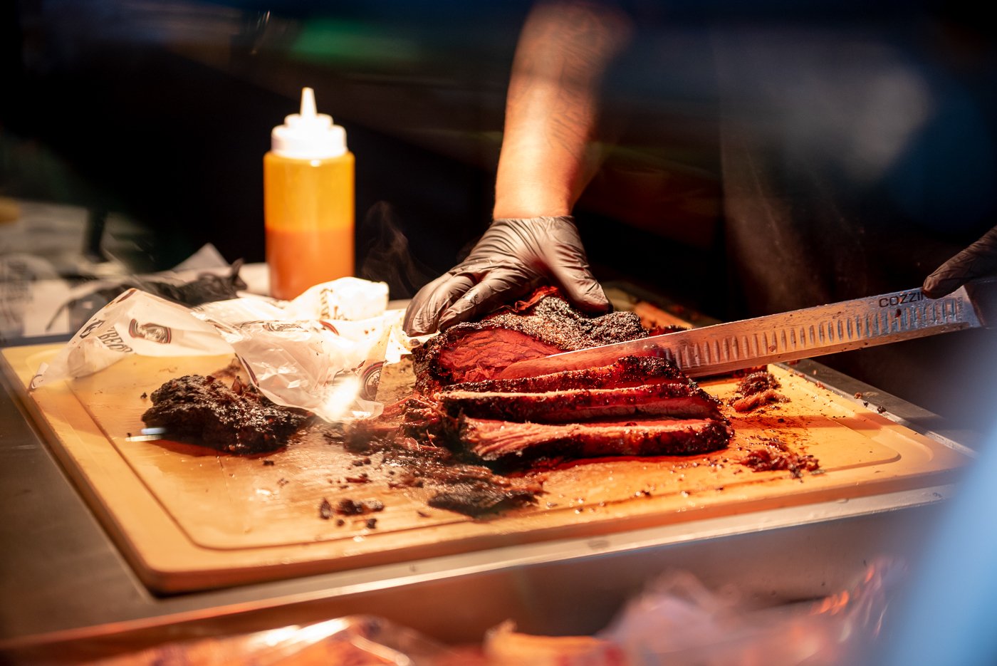 Chef slicing smoked brisket on a wooden cutting board with black gloves, with barbecue sauce bottle in the background.