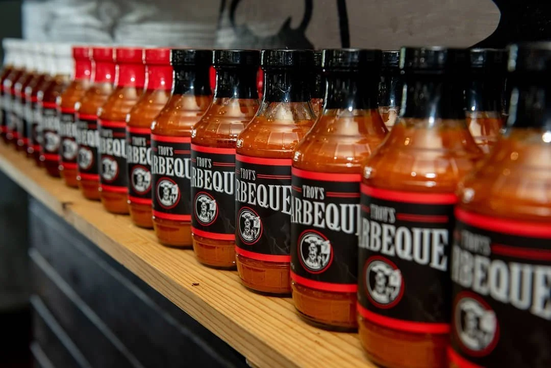Rows of barbecue sauce bottles labeled 'Troys Barbecue' on a wooden shelf.