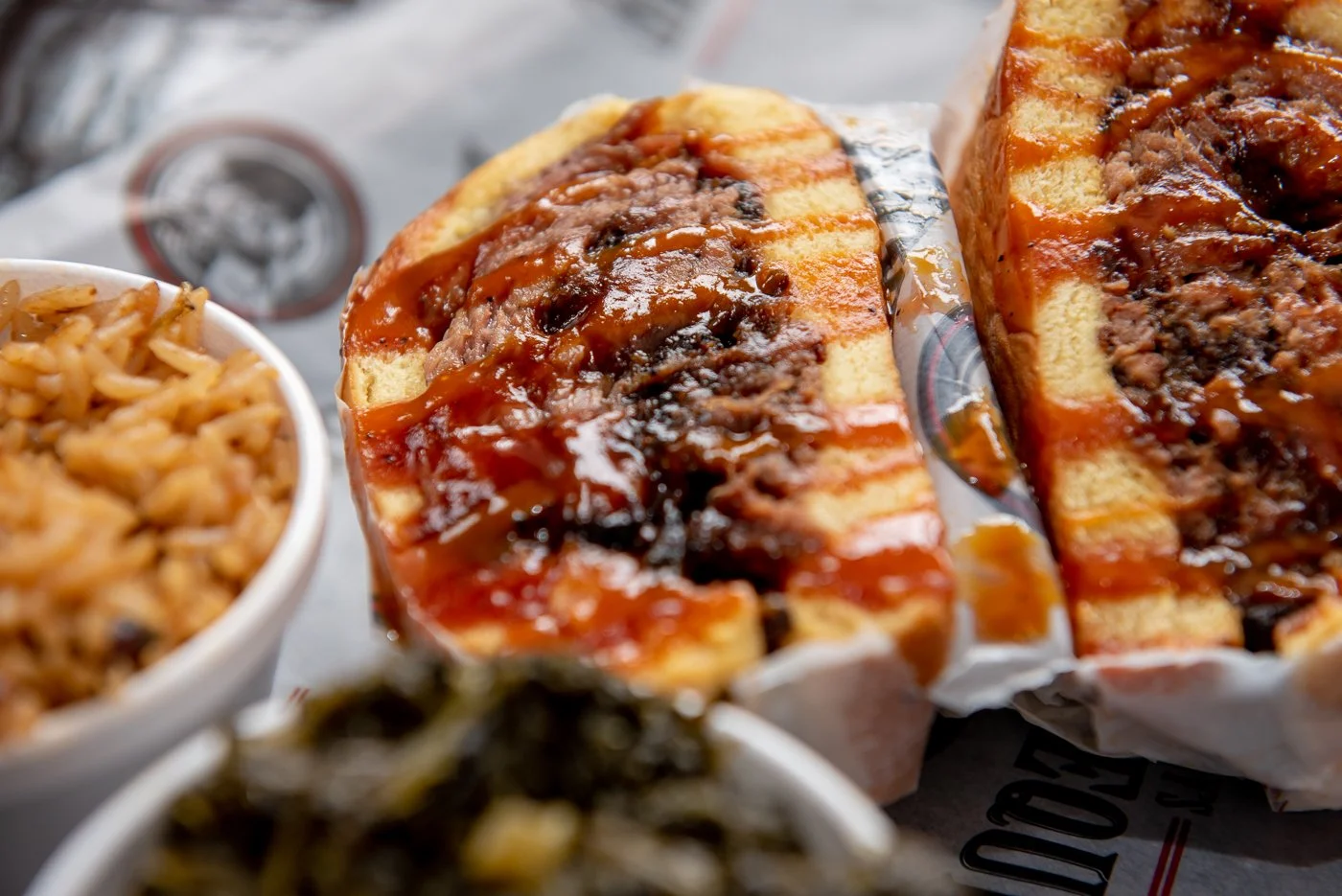 A sliced serving of beef brisket sandwich with barbecue sauce on a piece of parchment paper, with side dishes including coleslaw and pickles in bowls.