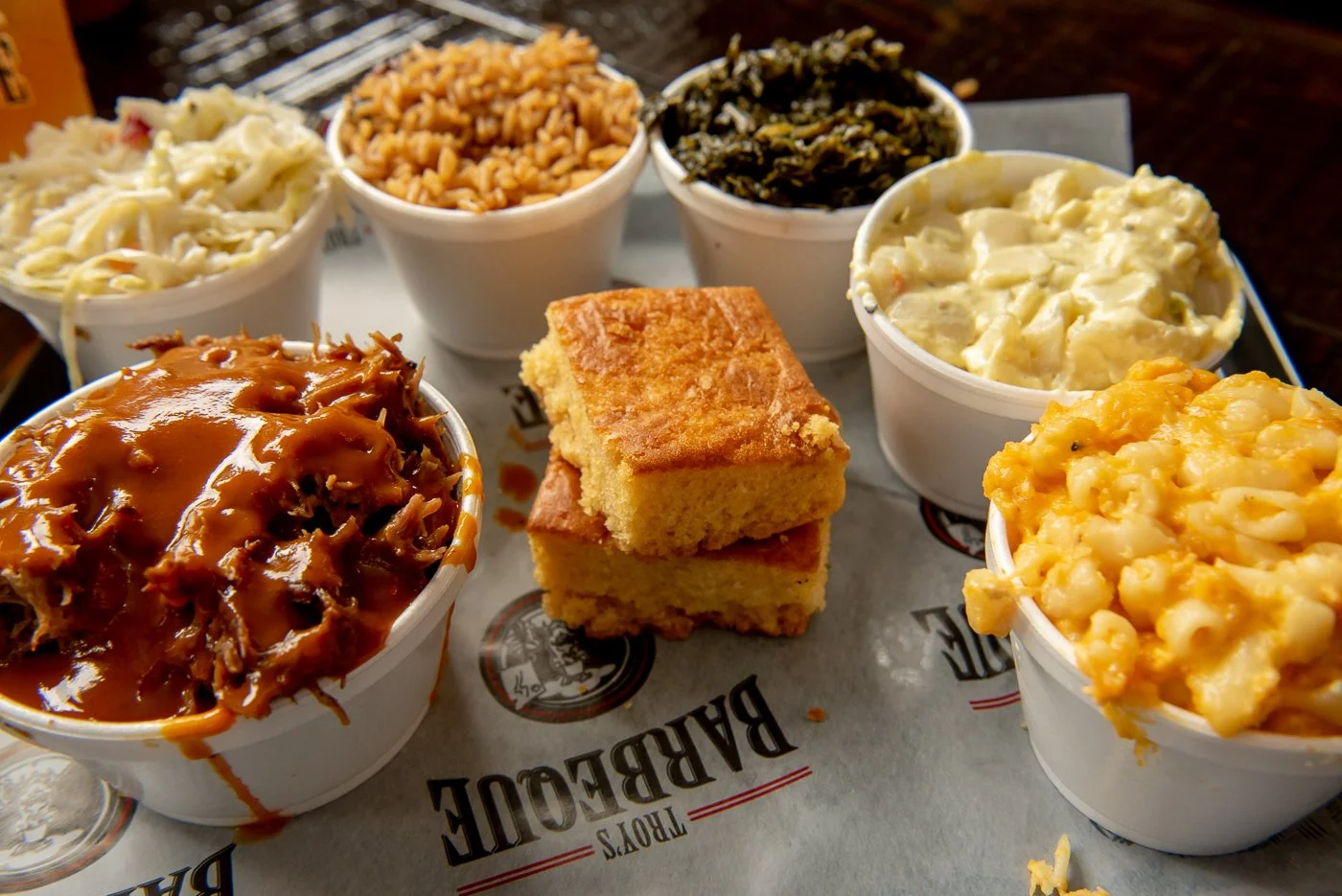 Various bowls of barbecue sides including baked mac and cheese, greens, BBQ baked beans, and potato salad, with a serving of barbecue meat and cornbread on a tray.