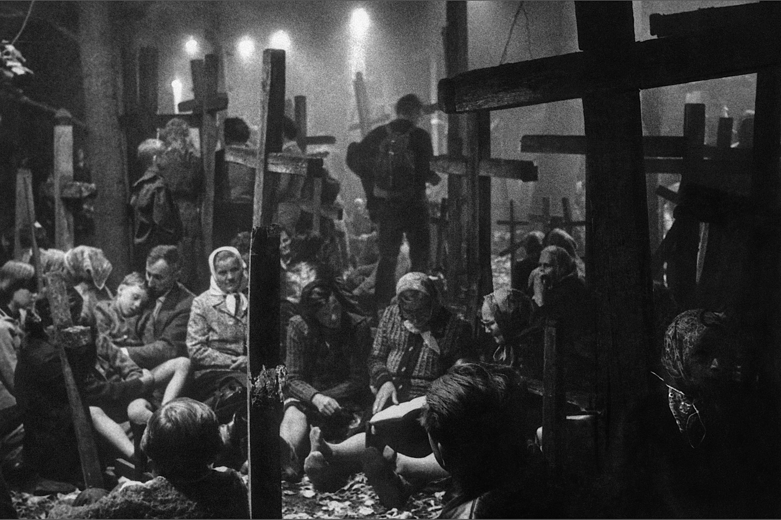 Black and white photo of people inside a concentration camp with wooden crosses and barbed wire, graves, and guards in the background.