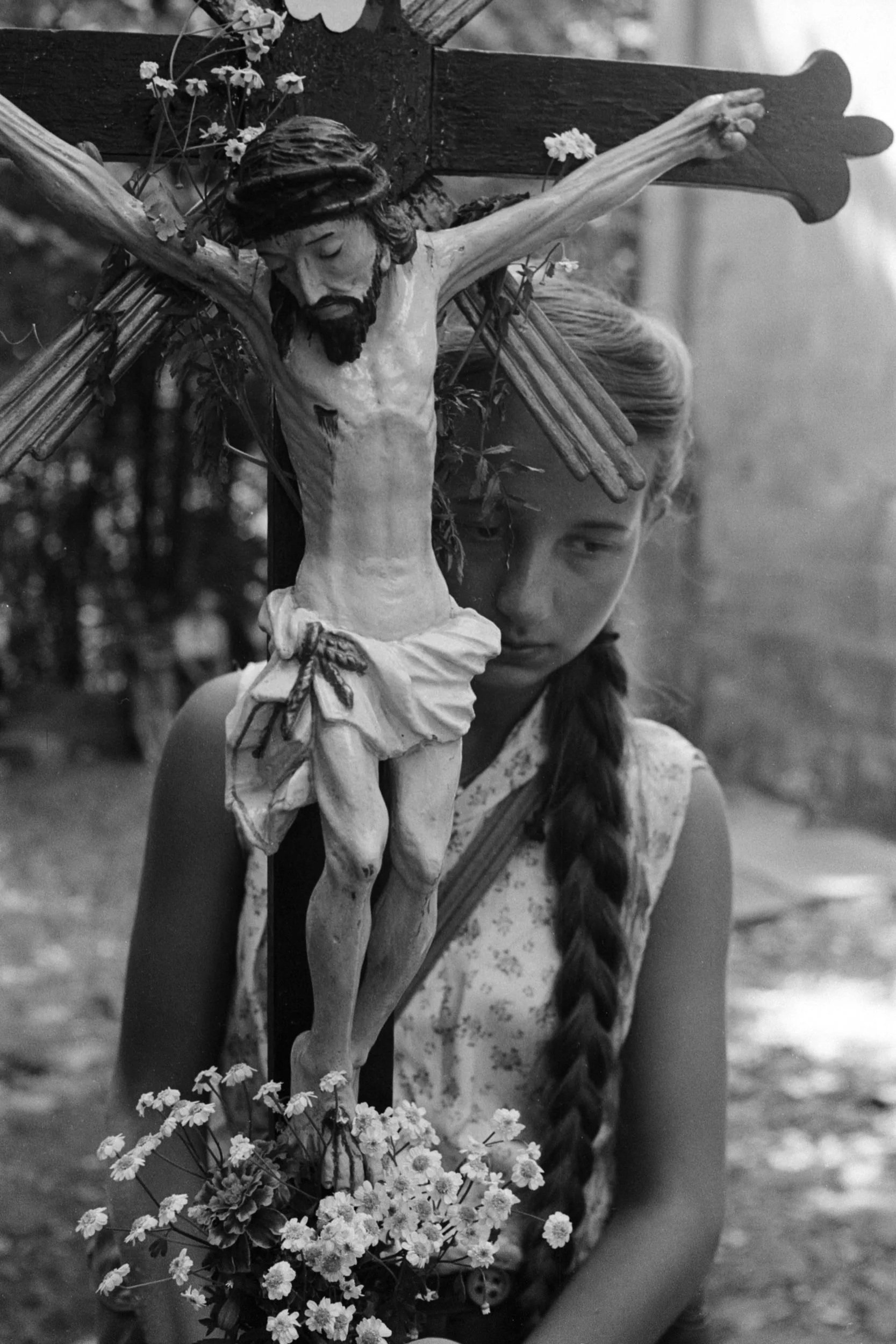 A girl with braided hair stands behind a crucifix with a detailed figure of Jesus Christ, holding a bouquet of flowers, in an outdoor setting.