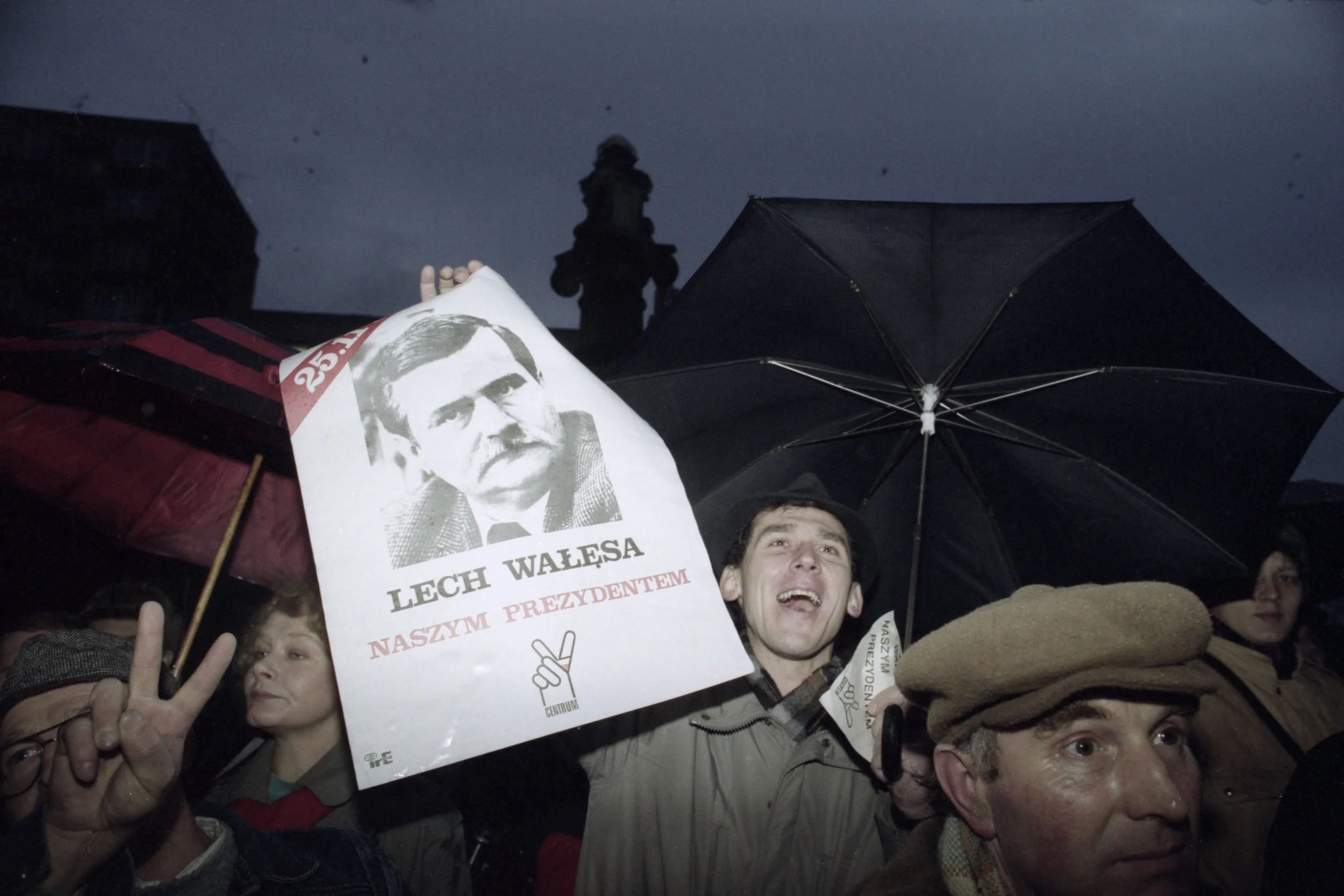 Protesters holding a portrait of Lech Walesa, with one person showing a peace sign and another holding an umbrella, during an outdoor protest in rainy weather.