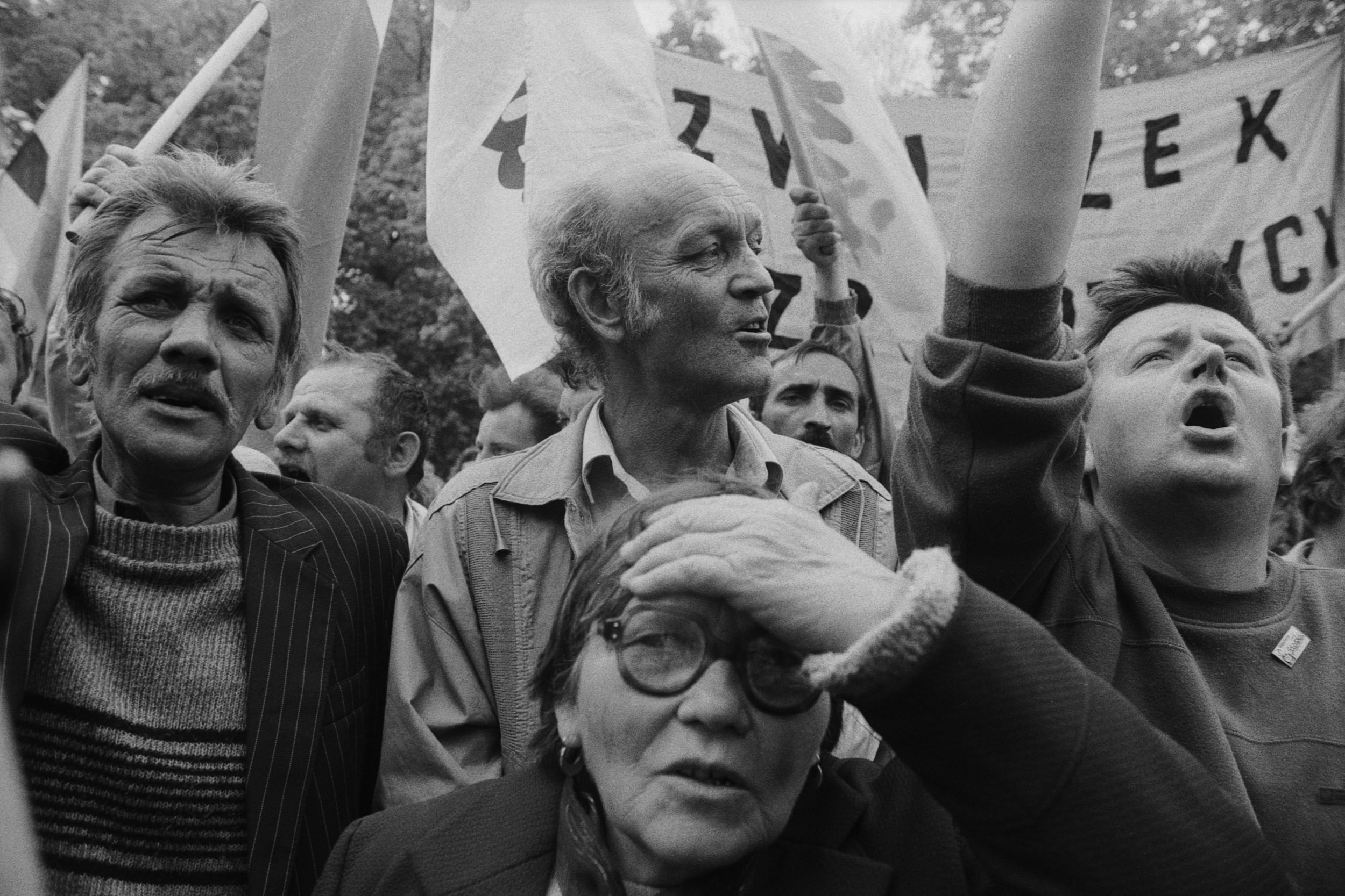 A black and white photo of a crowd protesting or rallying, holding flags and banners, with diverse expressions of concern and determination.