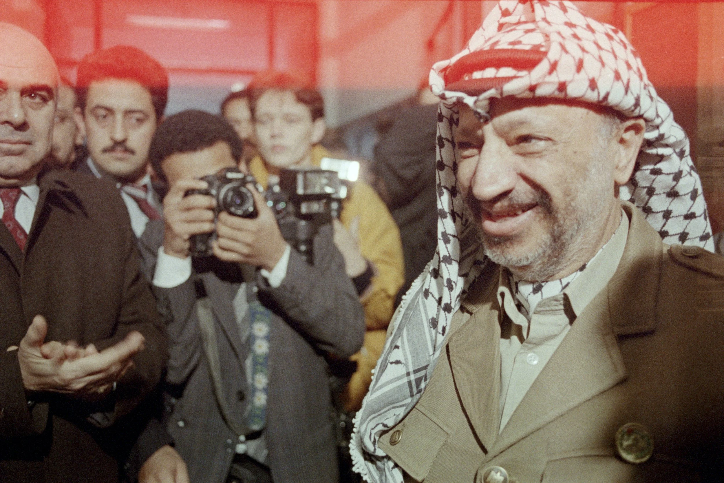 A crowd of people, including a man in a suit and tie, a man with a camera, and a man in military uniform wearing a checkered keffiyeh, smiling and engaging in conversation.