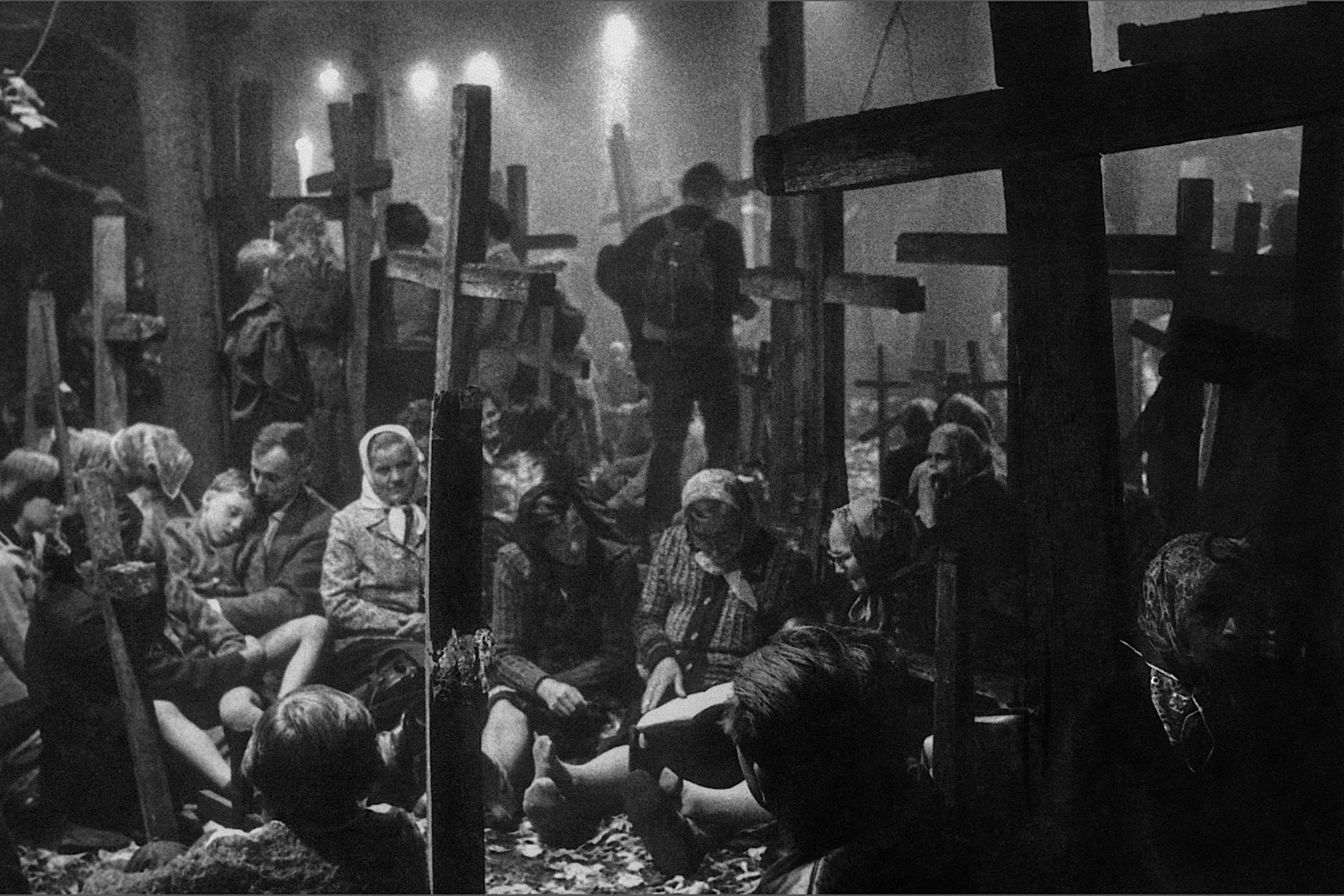 Black and white photograph of a somber scene with people sitting among wooden crosses in a cemetery, some wearing headscarves, others in casual clothing, with a few individuals standing in the background amidst faintly lit lights.