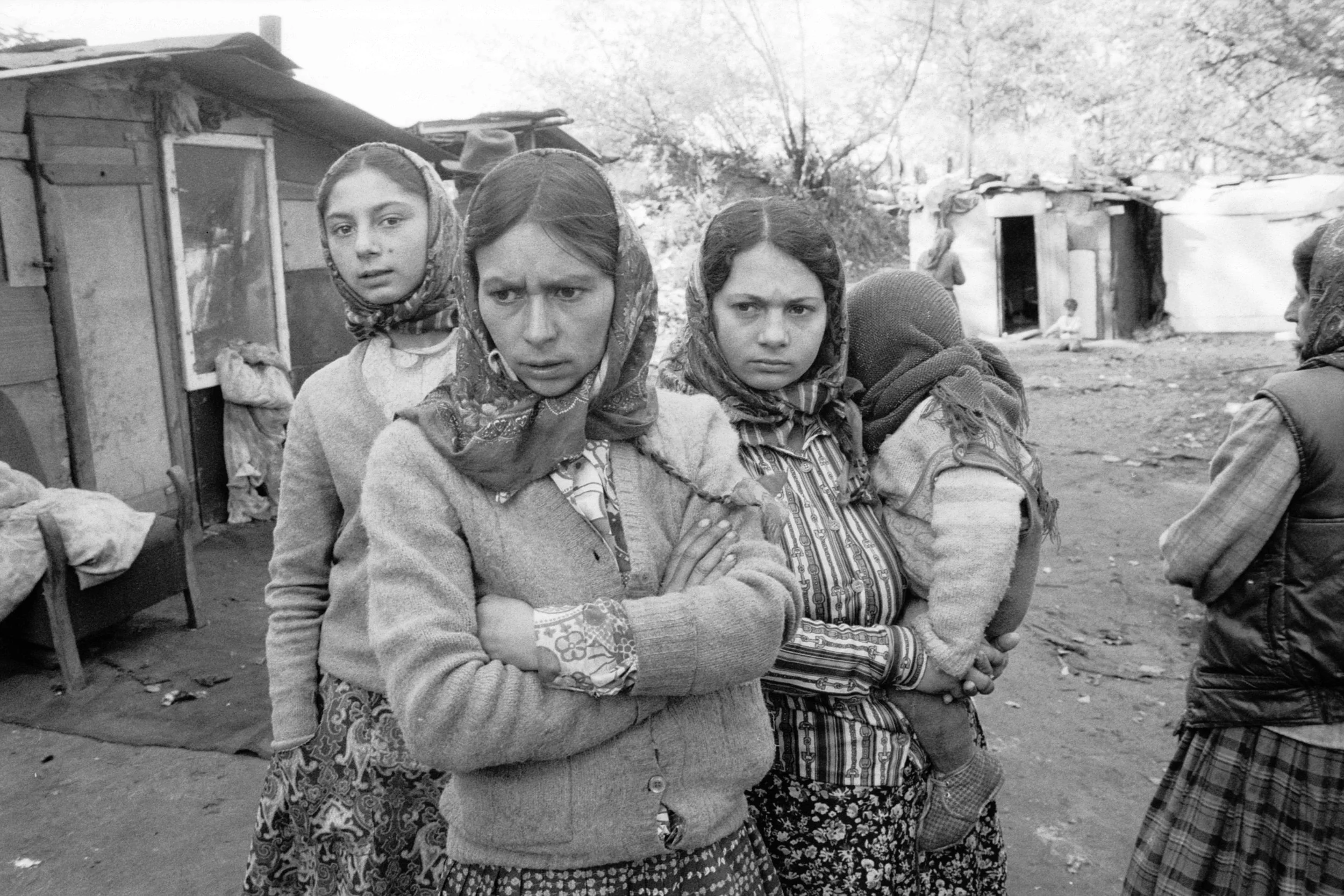 Four women with headscarves standing outside a makeshift home, looking serious or upset.